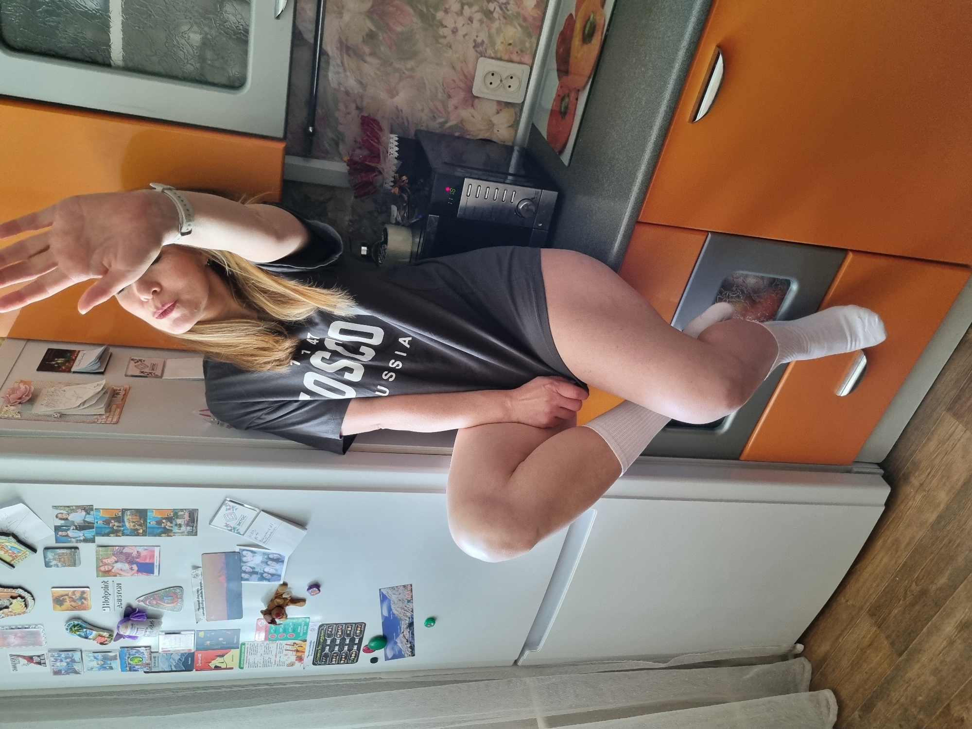 This image depicts an indoor scene in a kitchen within the city of Chaikovsky, Russia. A female individual with light brown hair is seated on a kitchen countertop. She is wearing a dark grey t-shirt with "OSCO RUSSIA" printed in white on the front and white ankle socks. Her right arm is raised, with her open palm facing the camera, and a light-colored watch is visible on her right wrist. Her left leg is bent, while her right leg hangs downward.
To the left of the individual is a white refrigerator adorned with numerous magnets, including photographs, illustrations, and one identifying "Hotpoint Ariston." A white sheer curtain is visible along the left edge of the frame. The floor is covered with wood-patterned laminate flooring.
The countertop on which the individual is seated is dark grey, and the base cabinets beneath it are orange with silver handles. A black microwave oven displaying "11:18" is situated on the counter to the right of the woman. Further right, a white electrical outlet is mounted on the wall, which features floral patterned wallpaper. A glass cutting board with an image of sliced bell peppers is also visible on the countertop.