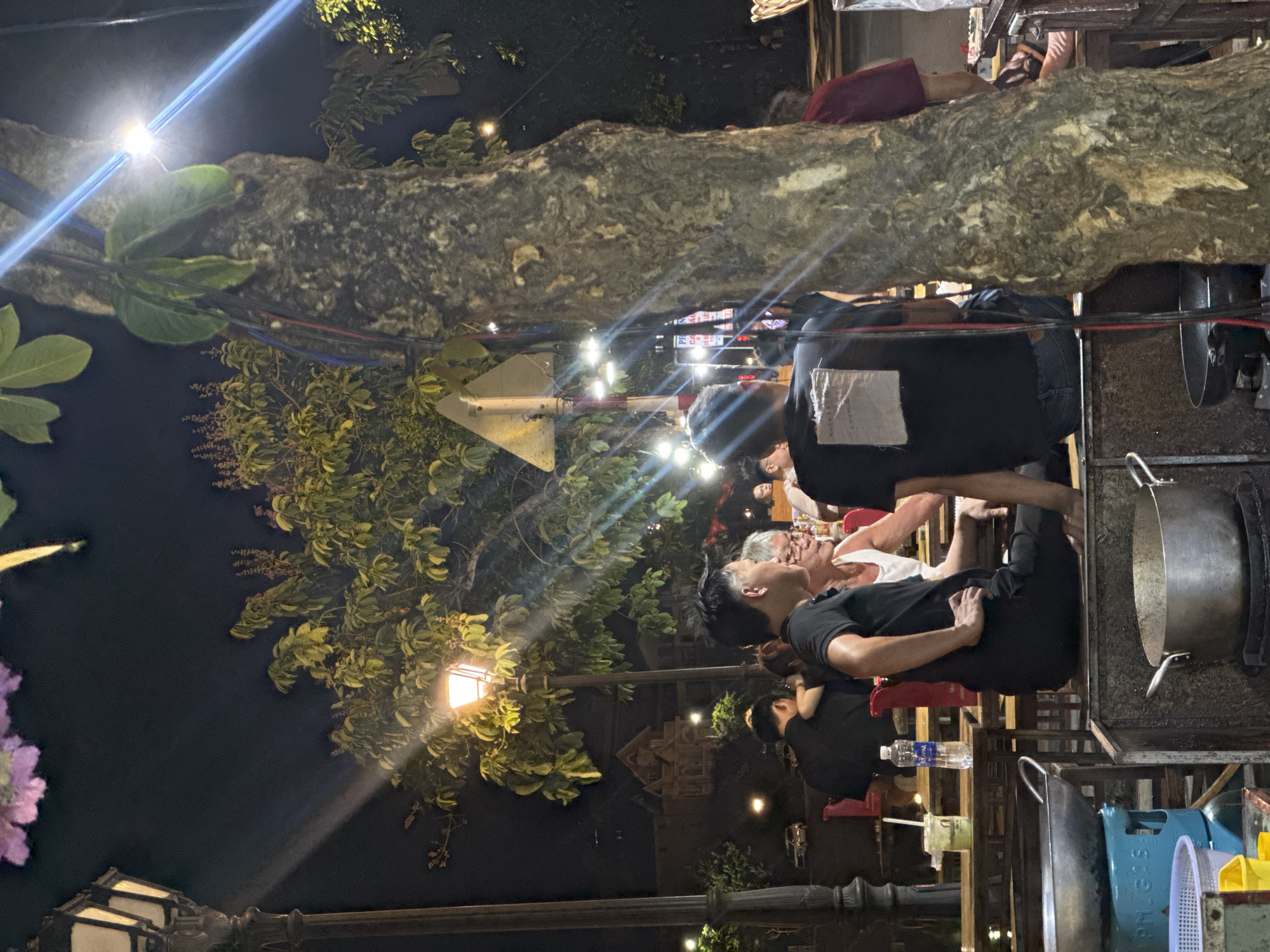 The image shows a night scene at a restaurant in Hội An Ward, Vietnam. Several people are seated at tables, some wearing black shirts and others in light-colored clothing. One individual is visible lying on a tree branch above the dining area. A large metal pot is situated on a cooking surface near the diners. A street lamp illuminates the scene with trees and foliage visible in the background. There is a street sign. Plastic storage containers are near the tables.