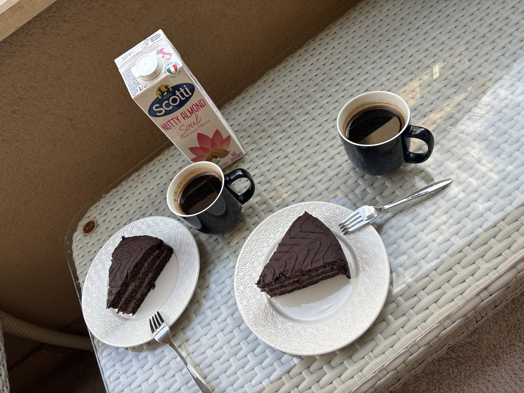 Two slices of chocolate cake and two cups of coffee are arranged on a white wicker table. The cake slices are on white plates with a decorative rim and a fork is placed next to each. A carton of Scotti Nutty Almond Soul plant-based beverage is positioned in the upper right corner of the table. The scene is outdoors, with natural light casting shadows on the table. The background appears to be a blurred outdoor environment, possibly a balcony or patio.