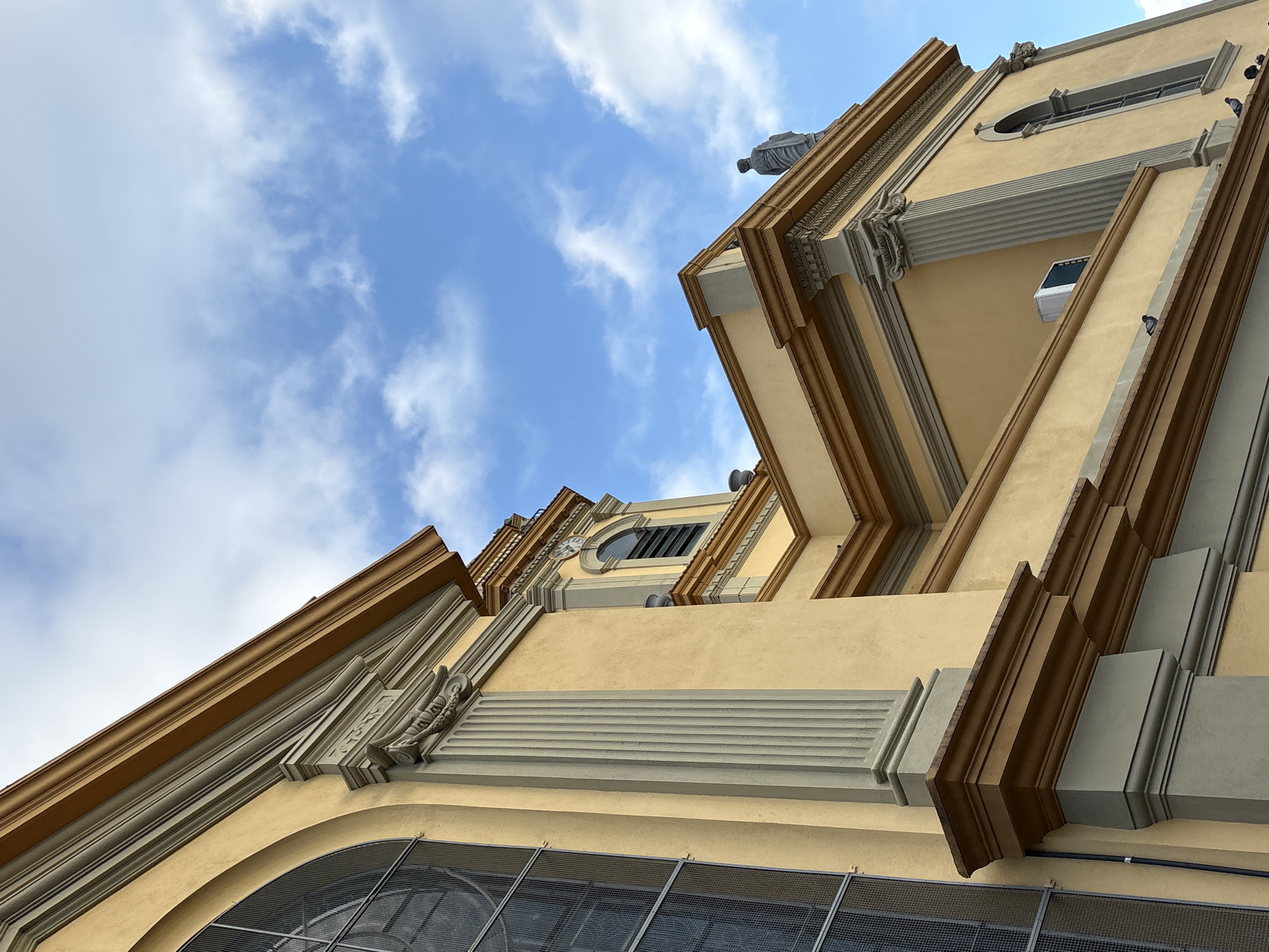 The image shows an upward angle view of the exterior of the Metropolitan Cathedral of São Paulo, Brazil. The building is made of stone and features a yellow facade with decorative elements. A large arched window with a metal grate is visible on the left side of the image. Further to the right, a section of the cathedral's tower rises, displaying a clock face and a small arched window. The roofline is adorned with cornices and sculptural details, and a statue of a figure stands on the upper edge of the building. The sky above is blue with scattered white clouds. Several pigeons are perched on ledges and architectural features of the building. An air conditioning unit is mounted on the exterior wall.
