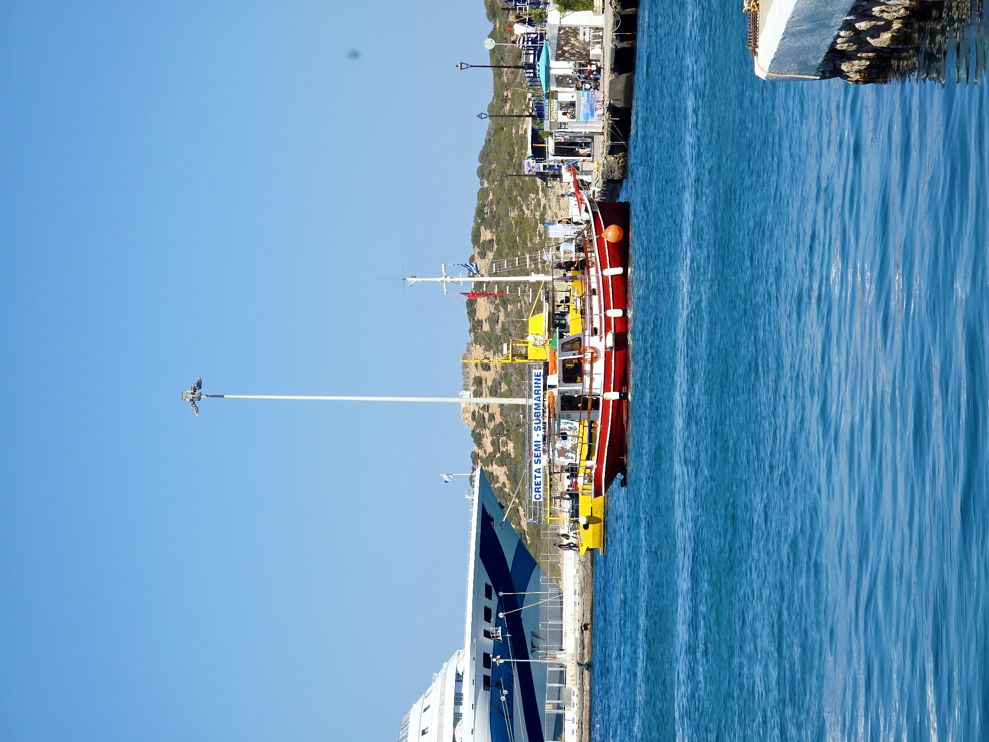 A bright, sunny day at a harbor in Agios Nikolaos Municipal Unit, Greece, is depicted in this image. The foreground is dominated by clear, blue water with gentle ripples. In the midground, several boats are docked. On the left, a large cruise ship with white and blue stripes is partially visible. To its right, a yellow boat is docked next to a red and white boat. The red and white boat has people on board and appears to be a tourist vessel, with the words "CRETA SEMI-SUBMARINE" visible on a banner. Further to the right, a pier with a canopy, tables, and people is visible. Beyond the harbor, a hilly, arid landscape with sparse vegetation and some buildings can be seen under a clear blue sky. A tall white lamppost stands prominently in the background.