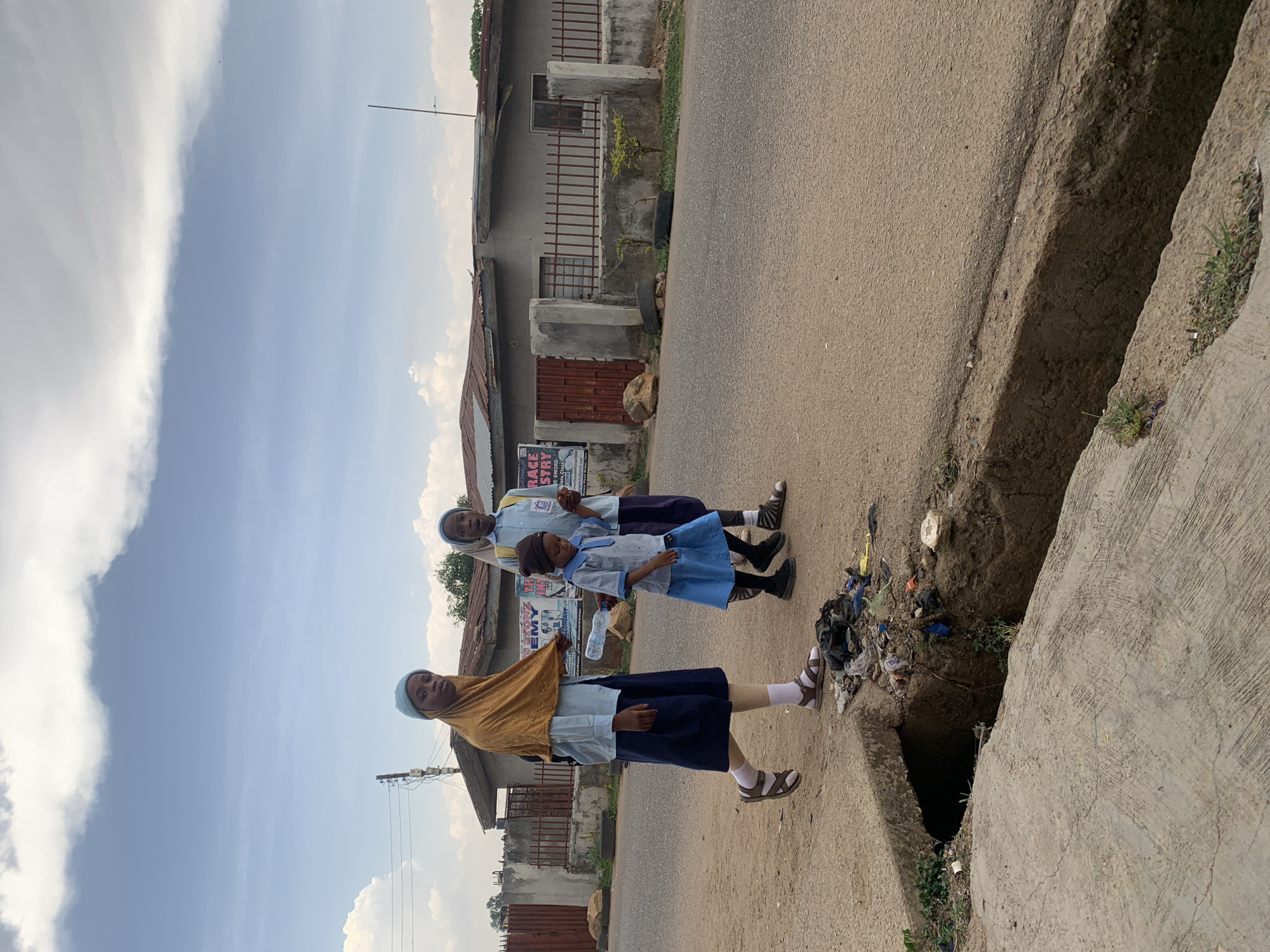 The image shows three schoolgirls in Gura Top, Nigeria, standing on the side of a road next to a drainage ditch. The girls are wearing school uniforms: light blue shirts, dark blue skirts, white socks, and sandals. The first girl on the left is wearing a light brown headscarf. The girl in the middle is holding a plastic bottle of water. The drainage ditch is filled with trash. Buildings are visible in the background, and the sky is partially cloudy.