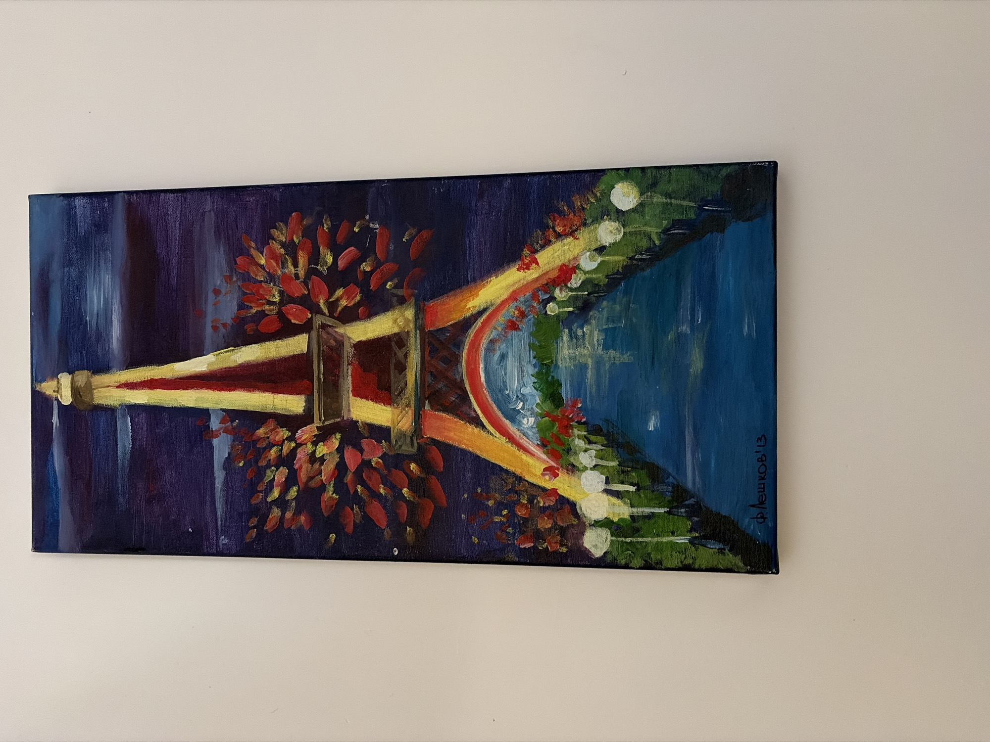 The image displays a vertical, rectangular oil painting of the Eiffel Tower at night. The tower is rendered in shades of yellow and red, illuminated against a dark blue, starry sky. Red and yellow foliage is depicted around the mid-section of the tower, and a rainbow arches over a blue body of water in the foreground. The water reflects the lights and colors of the tower and the surrounding environment. Along the banks of the water, there are rows of white, spherical lights and green foliage. The bottom left corner of the painting features a signature in dark red paint which reads "Флешков'13". The painting is hung on a plain, off-white wall. The location provided, Germasogeia Municipality, Cyprus, is not depicted in the painting itself.