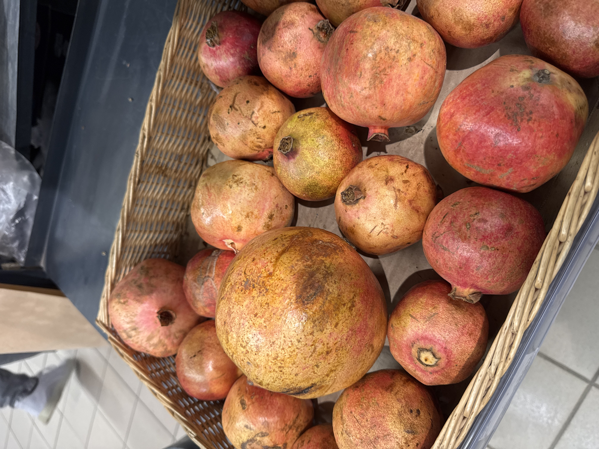 A close-up, high-angle shot shows a wicker basket filled with pomegranates. The pomegranates are round with textured skin that ranges in color from light orange to deep red, with some showing brown blemishes. They are arranged closely together, filling the basket. In the blurred background, a person's legs and feet are visible, wearing light-colored athletic shoes and grey pants, walking on a tiled floor. The floor tiles are a light beige color. The scene appears to be indoors, possibly a market or grocery store, in a location that has an agricultural background.