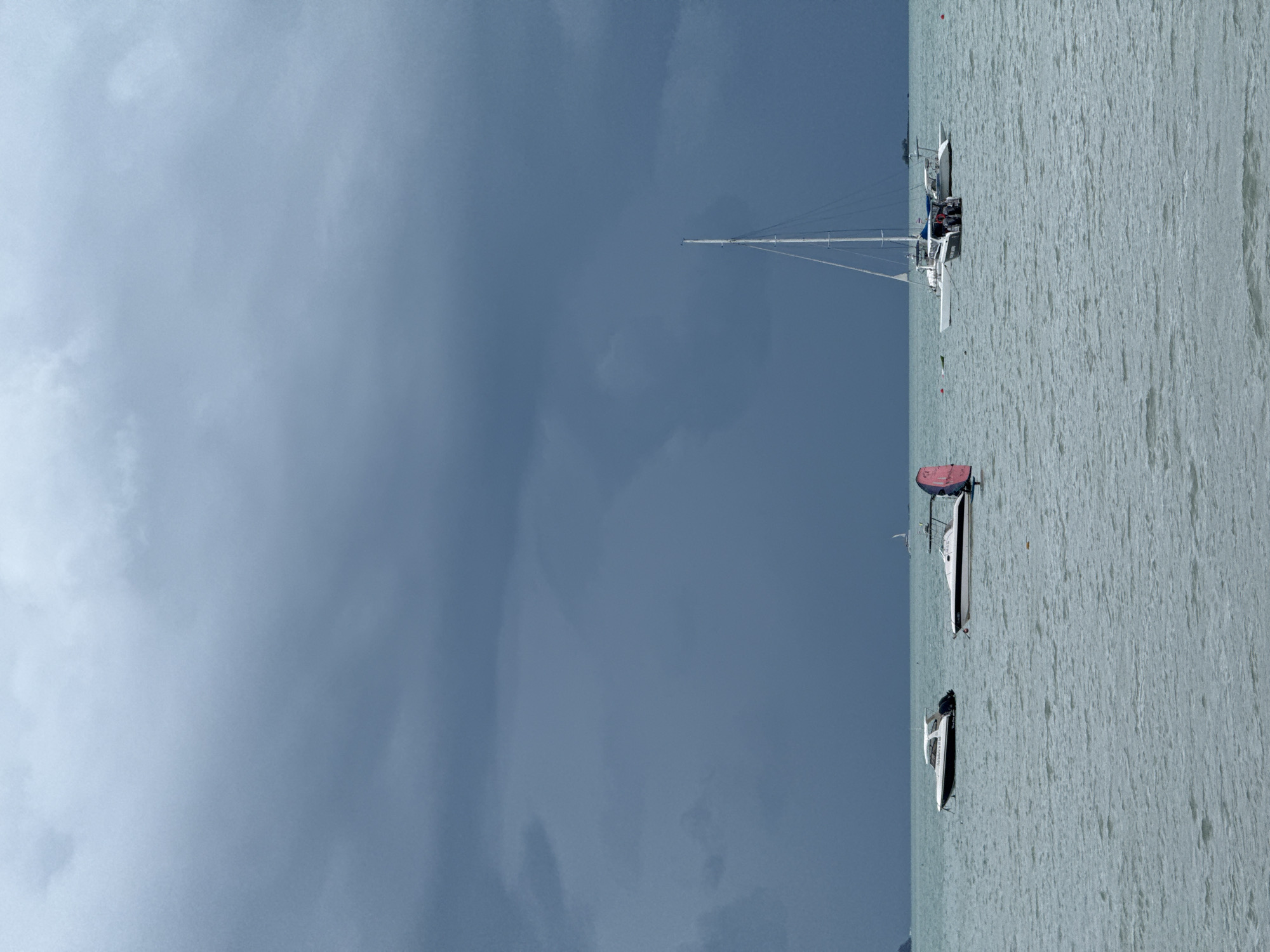 The image depicts a choppy sea under a dark, overcast sky. Several boats are visible on the water. In the foreground, there are two white speedboats, one with a black hull trim. To the right, a white catamaran with a tall mast is anchored, with a smaller boat in front of it. Further in the distance, a white sailboat is visible. The shoreline, or landmass, is only faintly visible on the far left. The scene appears to be a coastal area in Koh Samui District, Thailand, likely during a period of impending or recent rainfall, given the heavy cloud cover. No people are clearly visible on the boats.