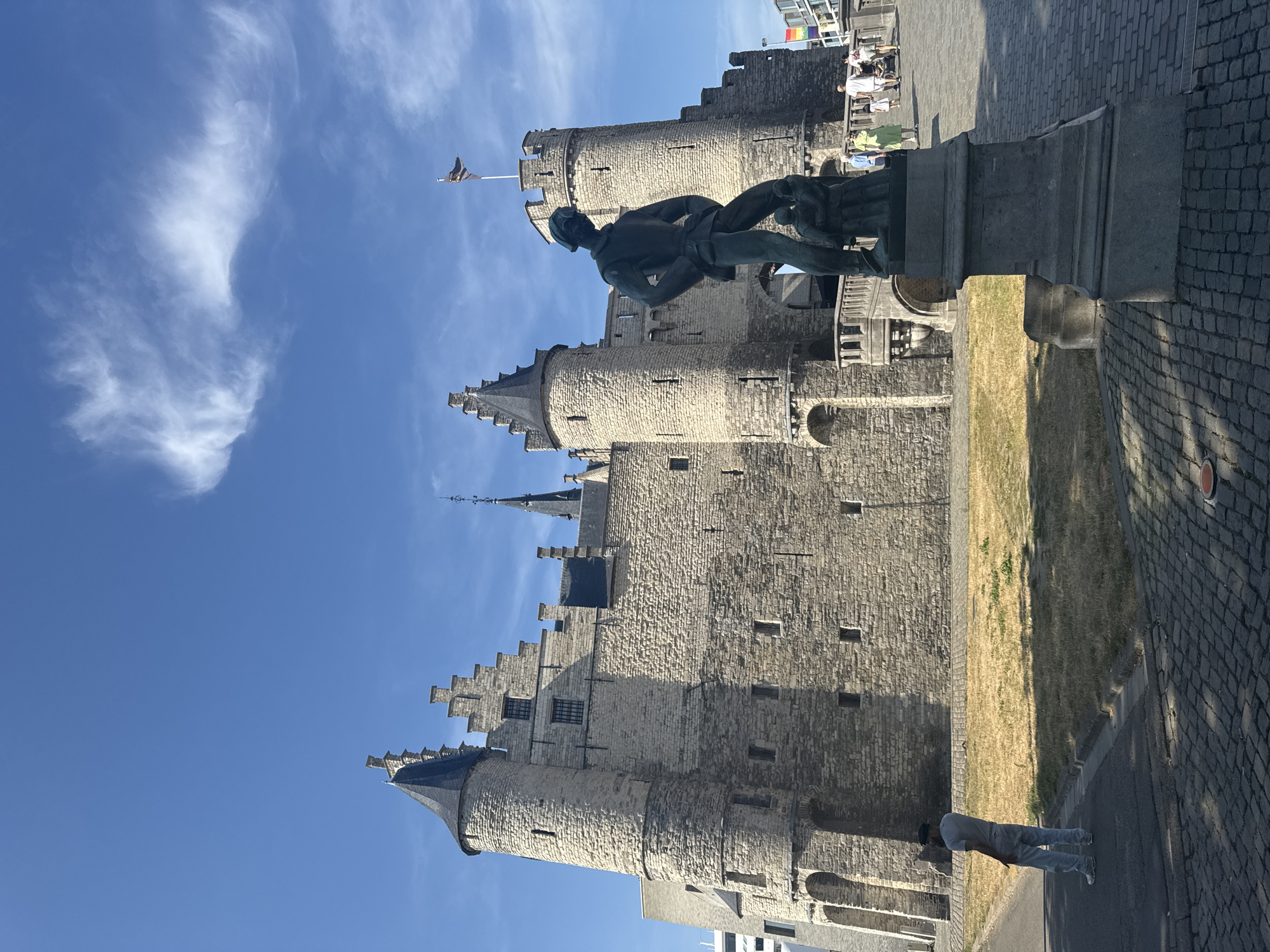 The image shows the Steen Castle in Antwerp, Belgium, on a clear, sunny day. In the foreground, a bronze statue depicts a man and two smaller figures. The statue stands on a stone pedestal. Behind the statue, the castle is visible, characterized by its stone walls, turrets, and stepped gables. A flag flies from one of the castle's towers. Several people are walking on a bridge leading towards the castle. A lone man stands on the paved area in front of the castle, facing away from the camera. The ground is paved with cobblestones. The environment is urban, with a hint of dry grass in front of the castle.