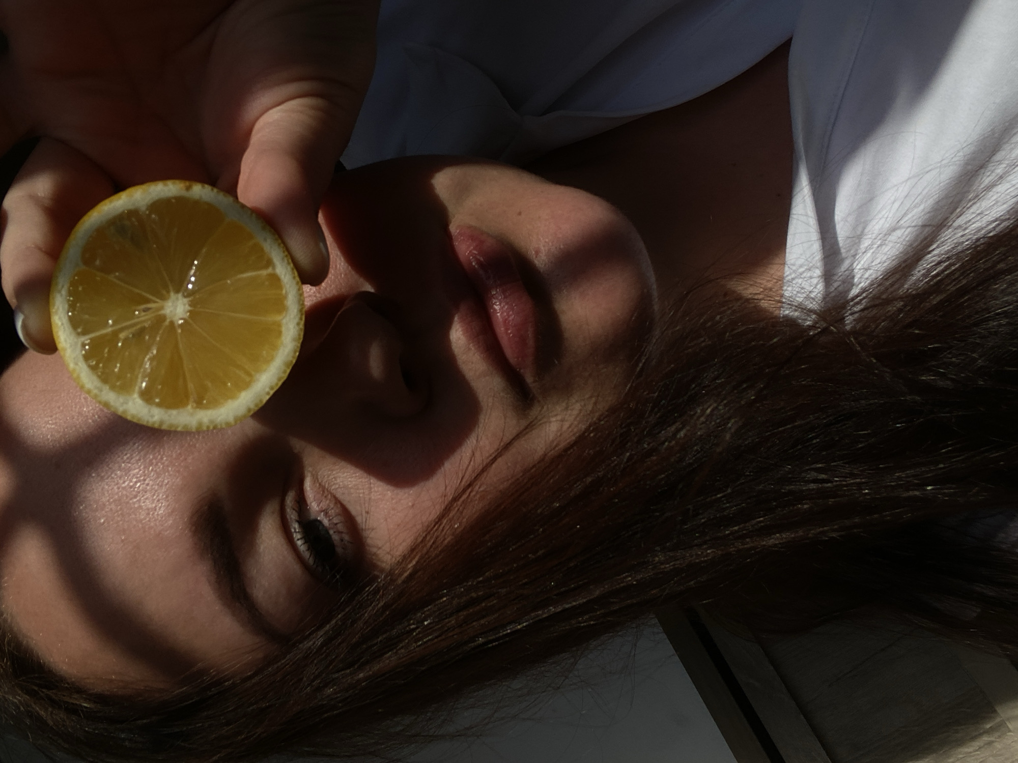 A woman with dark brown hair is lying on her back, wearing a white shirt. She is holding a slice of lemon in front of her left eye. The light source creates strong shadows on her face and hair. The background is blurred. Location information (Sochi, Russia) is not discernible from the image.