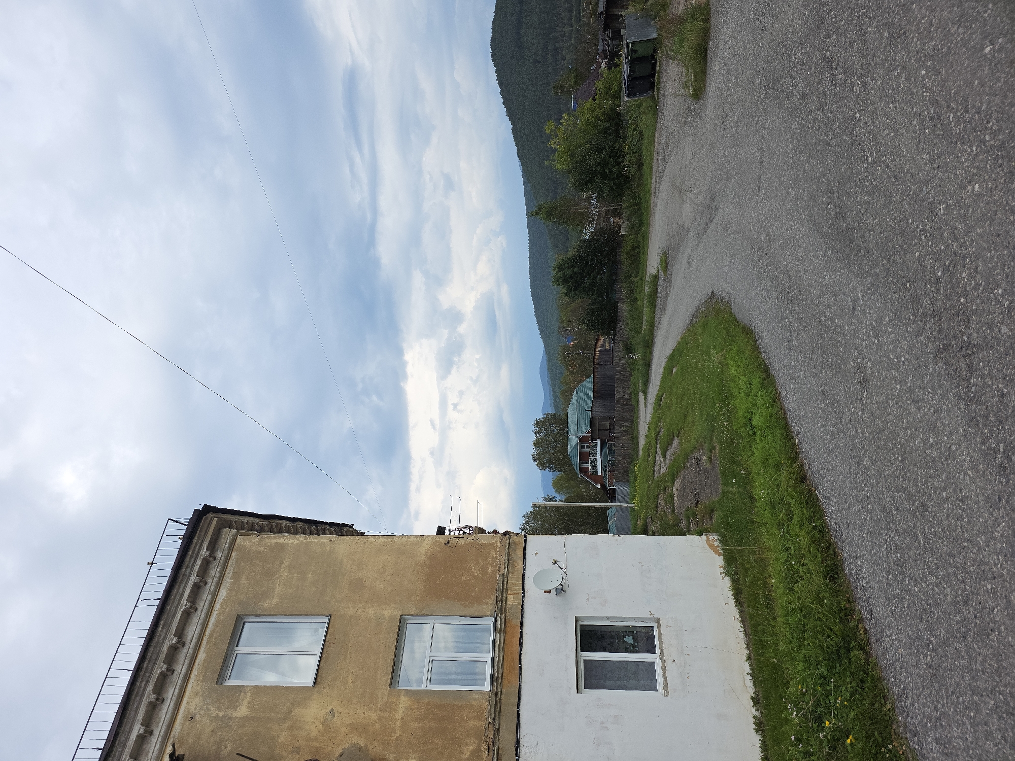 The image shows a curved asphalt road in a rural or semi-rural area. On the left, a two-story building with a weathered yellow facade and a white lower section is visible. This building has several windows, one with a satellite dish mounted on the wall below it. A metal railing runs along the top of the building. The road is bordered by grass and some small plants. In the background, there are several houses with pitched roofs, some surrounded by fences, and lush green trees. Beyond the houses, rolling hills covered in dense forest extend to the horizon under a cloudy sky. The scene suggests a quiet, somewhat undeveloped locality.
