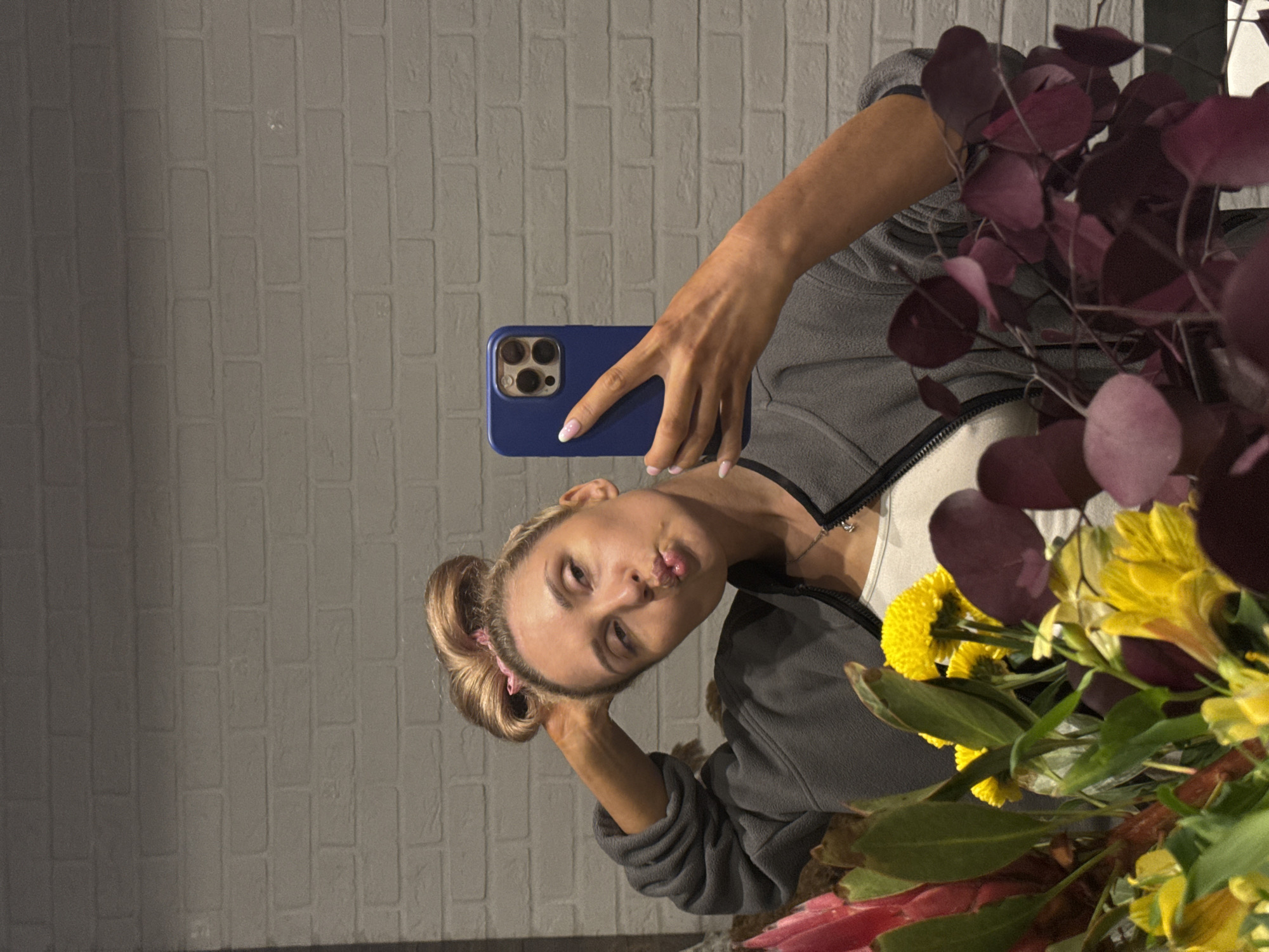 The image depicts an indoor scene in the city of Chaikovsky, Russia, featuring a female individual taking a selfie. The woman, with blonde hair styled in a high bun secured by a pink scrunchie, is wearing a grey fleece zip-up jacket over a white tank top and a silver necklace with a small pendant. Her fingernails are long and manicured with white tips. She holds a blue-cased smartphone (consistent with an iPhone Pro Max model) in her left hand, capturing her reflection. Her head is tilted to the left, and her lips are pursed in a "kissing" expression, while her right arm is bent with her hand resting near her head. The background is a light grey, brick-patterned wall. The lower foreground is filled with a dense floral arrangement comprising bright yellow spherical flowers, yellow trumpet-shaped blooms, a large pinkish-red tropical flower on the left, and dark red/purple circular leaves on the right, interspersed with green foliage.