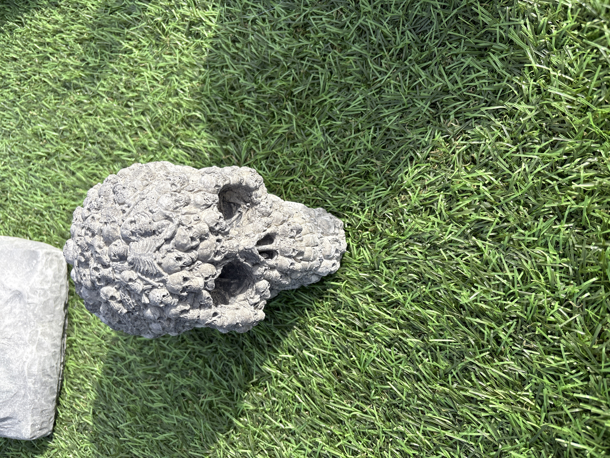 The image depicts a gray, skull-shaped object resting on artificial green turf. A rectangular gray object is partially visible on the left edge of the frame. The environment appears to be outdoors. The location is Chirk, United Kingdom.