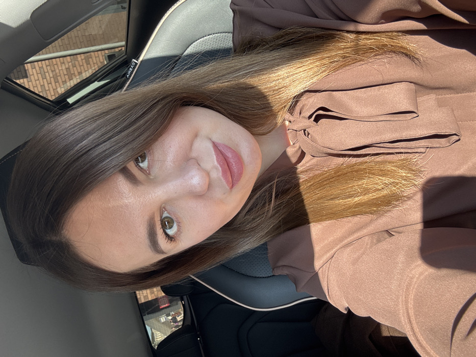 An adult female with light brown hair and light brown eyes, wearing a brown long-sleeved blouse with a pussy-bow necktie, is depicted in a selfie-style photograph. She is seated in the front passenger seat of a vehicle, identifiable by black and light-colored leather upholstery and an "AIRBAG" label visible on the pillar above the passenger seat. Strong sunlight enters from the right, casting distinct shadows on her hair, right cheek, and the front of her blouse. Through the rear passenger window on the left, an exterior scene is visible, showing a reddish-brown brick building and several indistinct figures of individuals walking. The scene is located in Likino-Dulyovo, Russia.