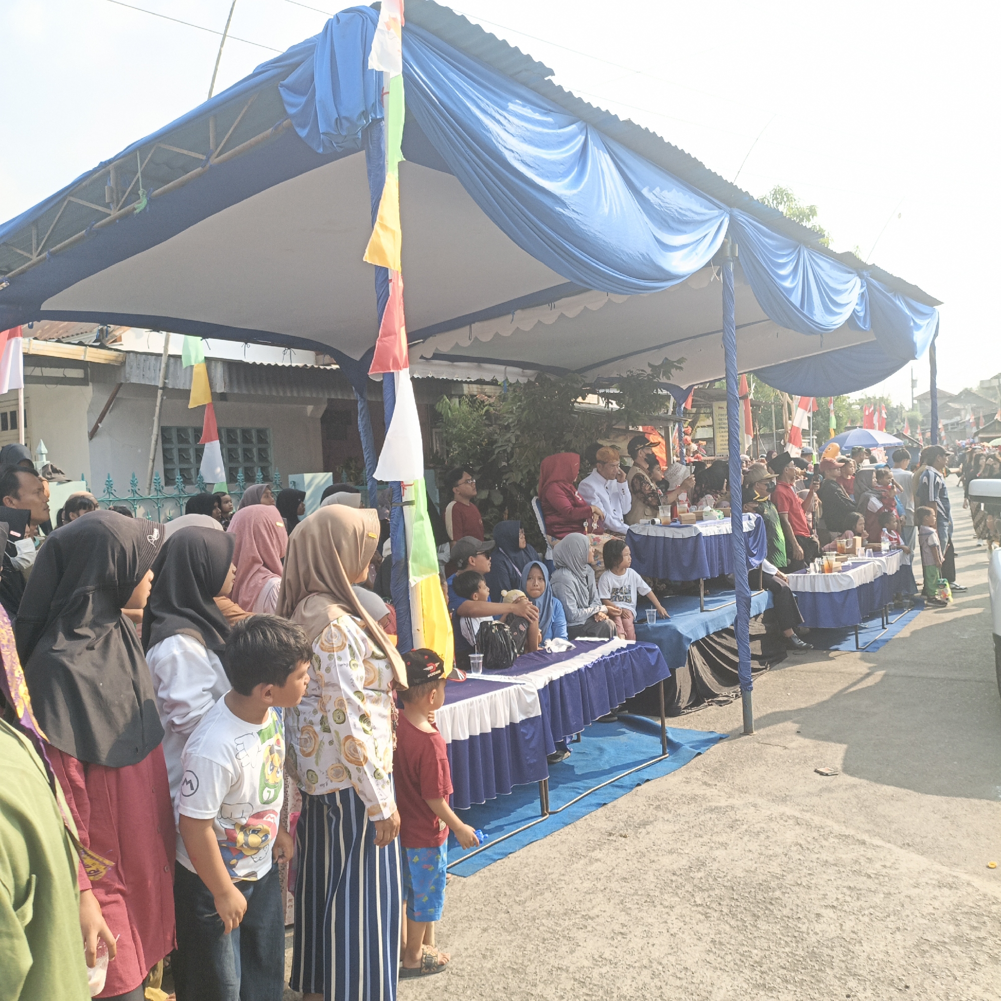 The image captures an outdoor event under a large blue and white tent, likely in Tanahbaya, Indonesia. A crowd of people, dressed in casual attire and headscarves, are gathered. Some are standing, while others are seated at tables covered with blue tablecloths. Many individuals appear to be watching an activity or performance happening to the right of the frame. The tent is supported by poles and decorated with Indonesian flags of red, white, green, and yellow. In the background, residential buildings with corrugated metal roofs are visible. The ground is paved and sunlight creates distinct shadows. Several individuals are holding cups or small objects. A child in a red shirt and blue shorts is standing in the foreground, looking to the right. Another child in a white shirt with a cartoon character is also prominent. The scene suggests a community gathering or celebration.