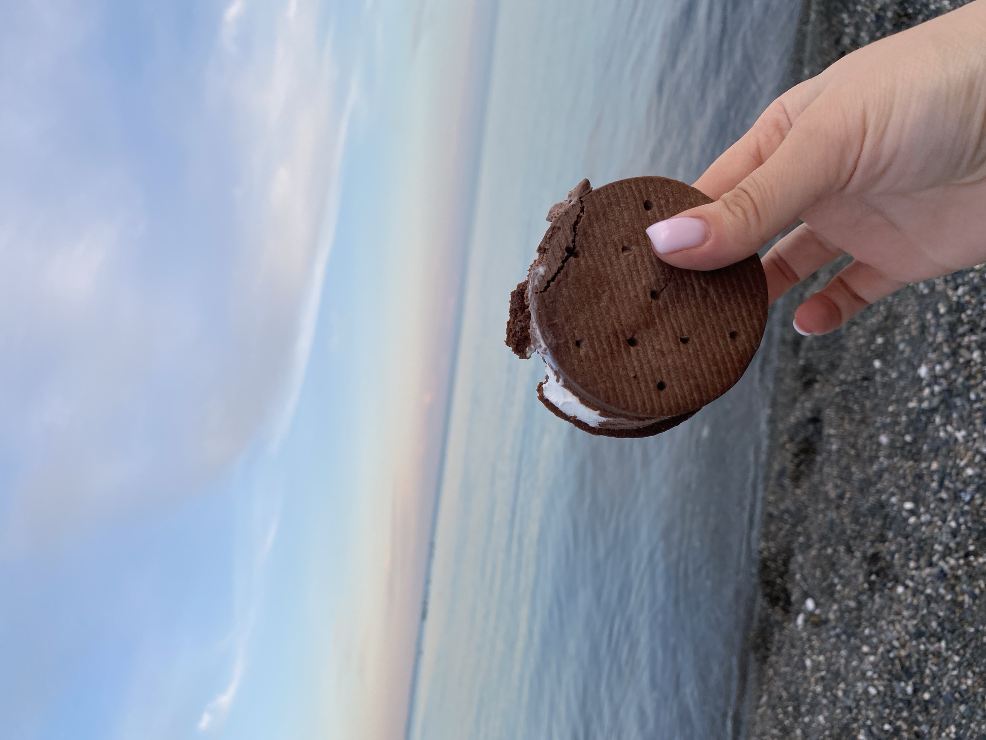 A hand holds a chocolate ice cream sandwich near a body of water. The sandwich has a bite taken out of it, revealing white ice cream. The hand has light skin with light pink painted nails. The background consists of the water and a sky with soft clouds. The ground in the foreground is a dark-colored pebble beach.