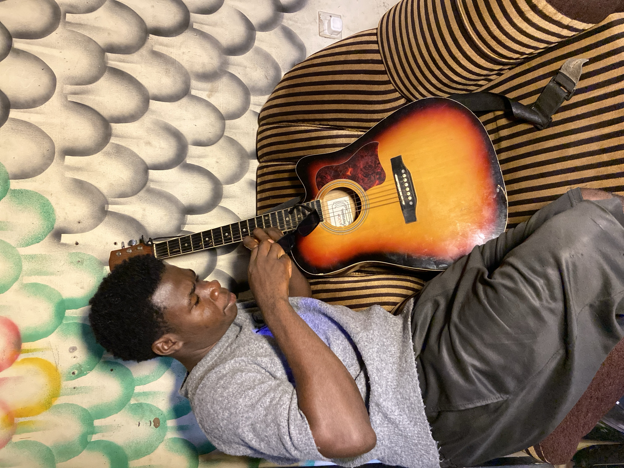 An overhead shot captures a young Black man lying down with an acoustic guitar in an indoor setting in Mararaba, Nigeria. The man, wearing a grey long-sleeved top and dark trousers, rests diagonally across the frame, with his head towards the bottom left. His eyes appear closed or partially closed, suggesting a moment of relaxation or contemplation, and his hands are positioned near the guitar's fretboard.
The acoustic guitar, featuring a sunburst finish (orange, yellow, and red hues) and a dreadnought body shape, is placed partially on the man's midsection and partly on a brown and tan/gold striped cushion or sofa arm. A black guitar strap is also visible. On the guitar's soundhole, a white label can be seen, partially displaying the words "FALCON" and "PROFESSIONAL".
The background consists of a wall adorned with a distinct painted pattern of overlapping light grey or white oval shapes, resembling scales. Towards the bottom left, this pattern incorporates splashes of green, red, and yellow circular accents. A white electrical outlet is visible on the wall in the upper right quadrant of the image. The scene suggests a tranquil moment of leisure.