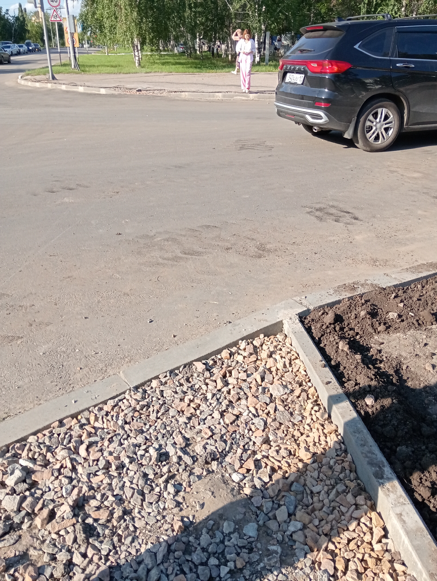 The image captures an outdoor scene on a sunny day, likely in a city environment. In the foreground, a newly constructed curb divides an area filled with loose gravel from a pile of dark soil. The asphalt road surface extends into the midground, showing some wear and tear with darker patches. In the background, a black SUV is parked on the road, facing away from the camera. To the left of the SUV, two individuals are standing on a grassy area adjacent to the road. One individual, appearing to be a woman, is wearing a white top and pink trousers and is looking to her left. A traffic light and various road signs are visible further down the road. The overall impression is of urban infrastructure undergoing or recently having undergone some form of construction or maintenance work, possibly near a pedestrian walkway or green space. The license plate of the SUV appears to be "S424JU 10."