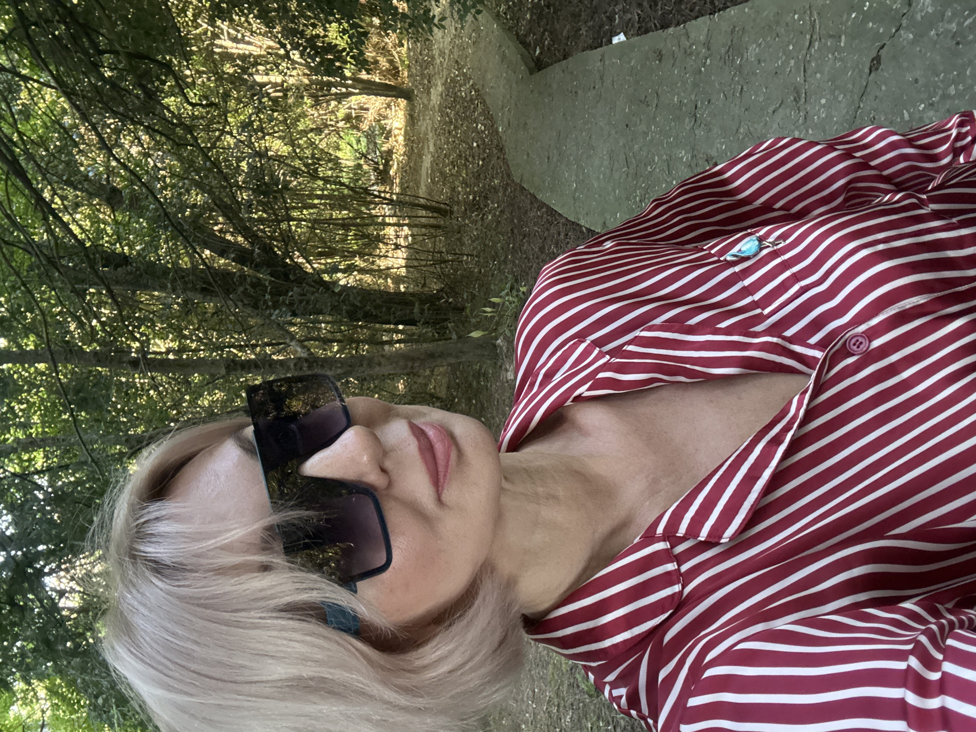 The media file presents a selfie-style photograph taken in a rotated portrait orientation, featuring a person outdoors in a wooded area of Sochi, Russia. The individual, likely female, is positioned on the right side of the rotated frame, with her head resting towards the left. She has short, light blonde hair and wears prominent, dark rectangular sunglasses with a subtle blue accent on the frames. Her lips are visible, adorned with light pink lipstick.
She is dressed in a red and white striped button-up shirt, which features a light blue bird-shaped brooch pinned to the chest pocket. The shirt's collar is slightly open.
The background, occupying the left side of the rotated frame, depicts a sun-dappled natural environment. Dense green foliage and tree branches are visible, with bright sunlight filtering through the leaves, suggesting a clear daytime setting. A dirt path covered with fallen leaves winds through the trees, and a strip of concrete or paved ground is visible towards the top right edge of the rotated frame. The image conveys a casual and relaxed moment in a natural park or forest setting.