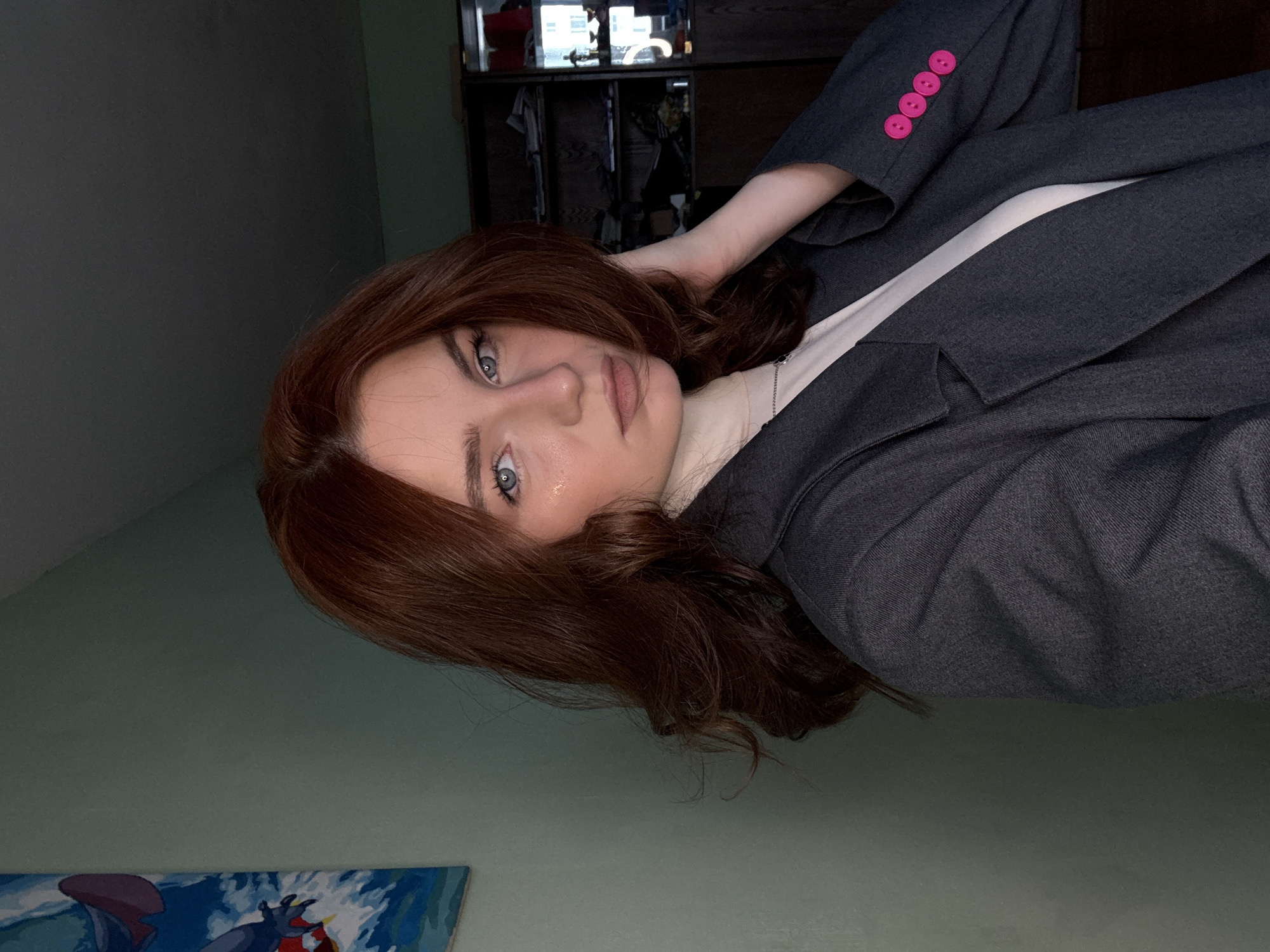 A woman with auburn hair and blue eyes is wearing a gray blazer with pink buttons and a white shirt. She is posing in front of a light green wall with a partial painting of blue and red objects in the bottom left corner. In the background, there is a dark wooden cabinet with items inside. The location is Unknown, Unknown.
