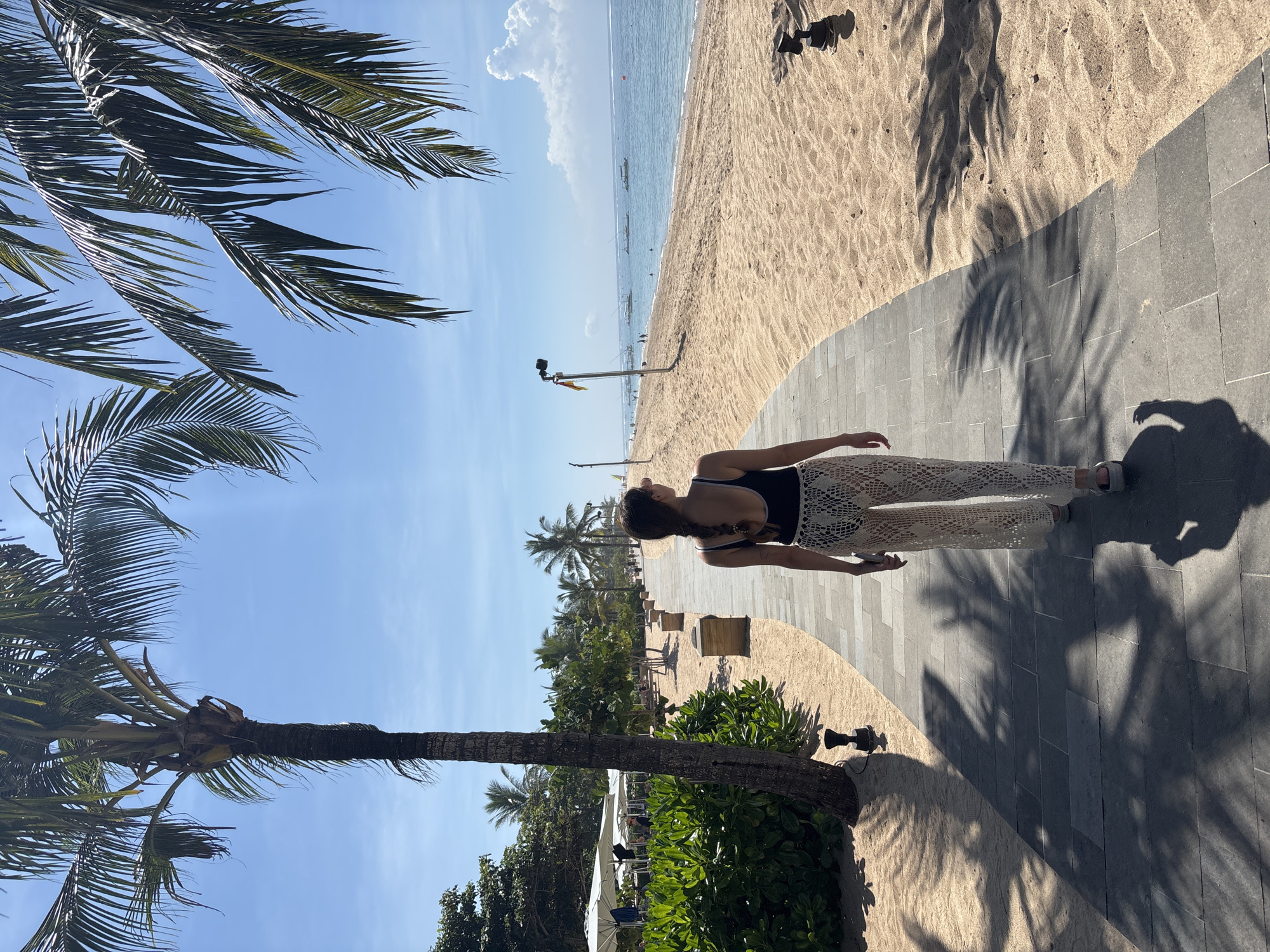 A medium shot captures a woman in Kuta Selatan, Indonesia, walking on a paved path adjacent to a sandy beach. She wears a black bathing suit top and crocheted pants. To her left, a palm tree extends into a blue sky. The beach and ocean stretch out in the background, and additional palm trees line the shore. The time of day is likely midday, indicated by the harsh shadows.