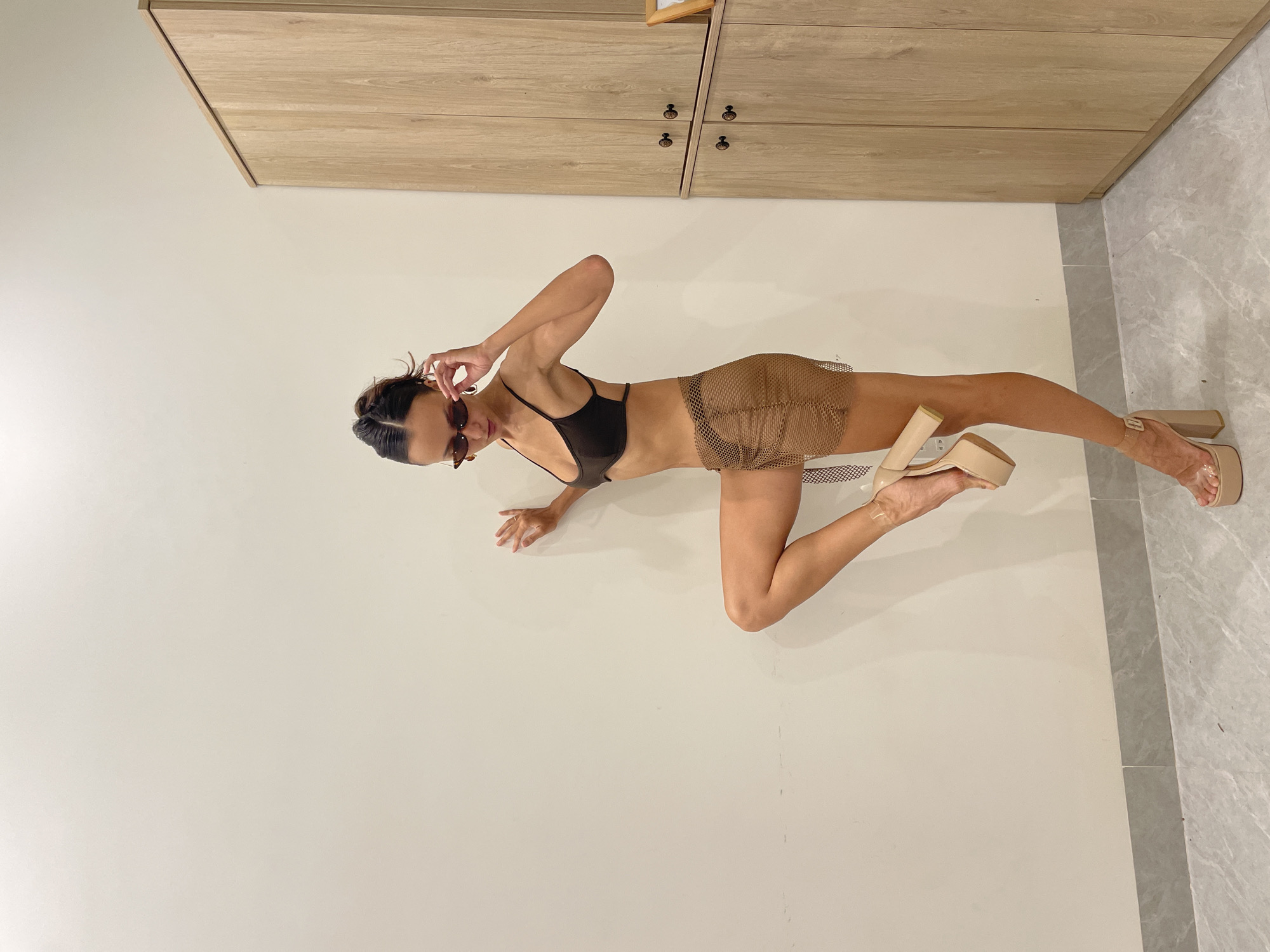 A full-shot image captures a woman striking a dynamic pose indoors, likely in Mũi Né, Vietnam. The woman, with dark hair pulled back, is seen crouching or leaning forward on a light grey tiled floor. She supports herself with her left hand on the floor and her right knee bent, while her right leg is extended backward with the foot planted, and her left foot is lifted. She wears a dark brown or black bikini-style top and a sheer, light brown, netted mini skirt. Dark sunglasses rest on her nose, and her right hand is raised to her face, seemingly adjusting them. Both feet are adorned with light beige or nude-colored platform high heels with clear straps.
The background consists of a plain, light-colored wall. Above the wall, along the top of the frame, are light wooden cabinets or drawers with dark, round handles, adding a warm, natural contrast to the minimalist setting. The lighting appears even and soft, suggesting an indoor environment. No text is visible in the image. The overall scene conveys a sense of fashion and confident posing.