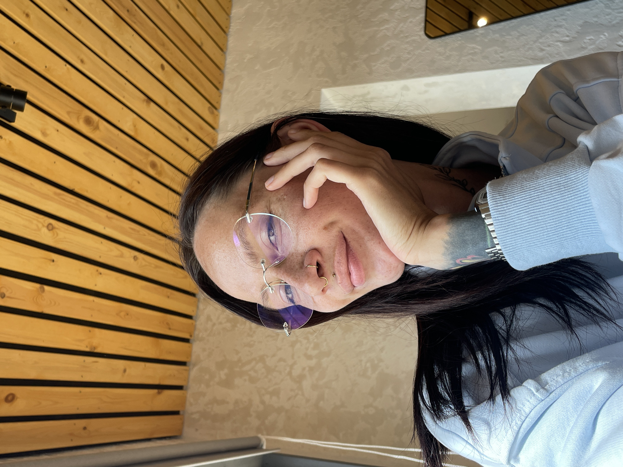 An indoor, eye-level portrait of a young adult woman is captured in Oktyabrsky, Russia. The woman, with long dark hair, gazes directly at the camera with a subtle smile, her left hand supporting her chin. She wears rimless glasses with gold-toned hardware and lenses exhibiting a faint purple reflection. Her face displays visible skin texture and freckles. She has a gold-toned horseshoe-shaped septum ring and a small gold-toned stud in her philtrum. A dark tattoo is visible on her left forearm, along with a silver-toned metal watch. She is dressed in a light blue hooded sweatshirt.
The background features a distinctive ceiling composed of parallel wooden planks separated by dark strips, angled diagonally across the upper portion of the frame. A black spotlight fixture is mounted on this ceiling in the upper right. The wall behind the woman is a light, textured surface. To her left, a vertical white element, possibly a window frame or blind cord, is partially visible. To her right, a portion of a rectangular mirror with a gold-toned frame reflects the wooden ceiling and an internal light source. The overall lighting suggests a bright, possibly naturally lit environment combined with artificial fixtures.