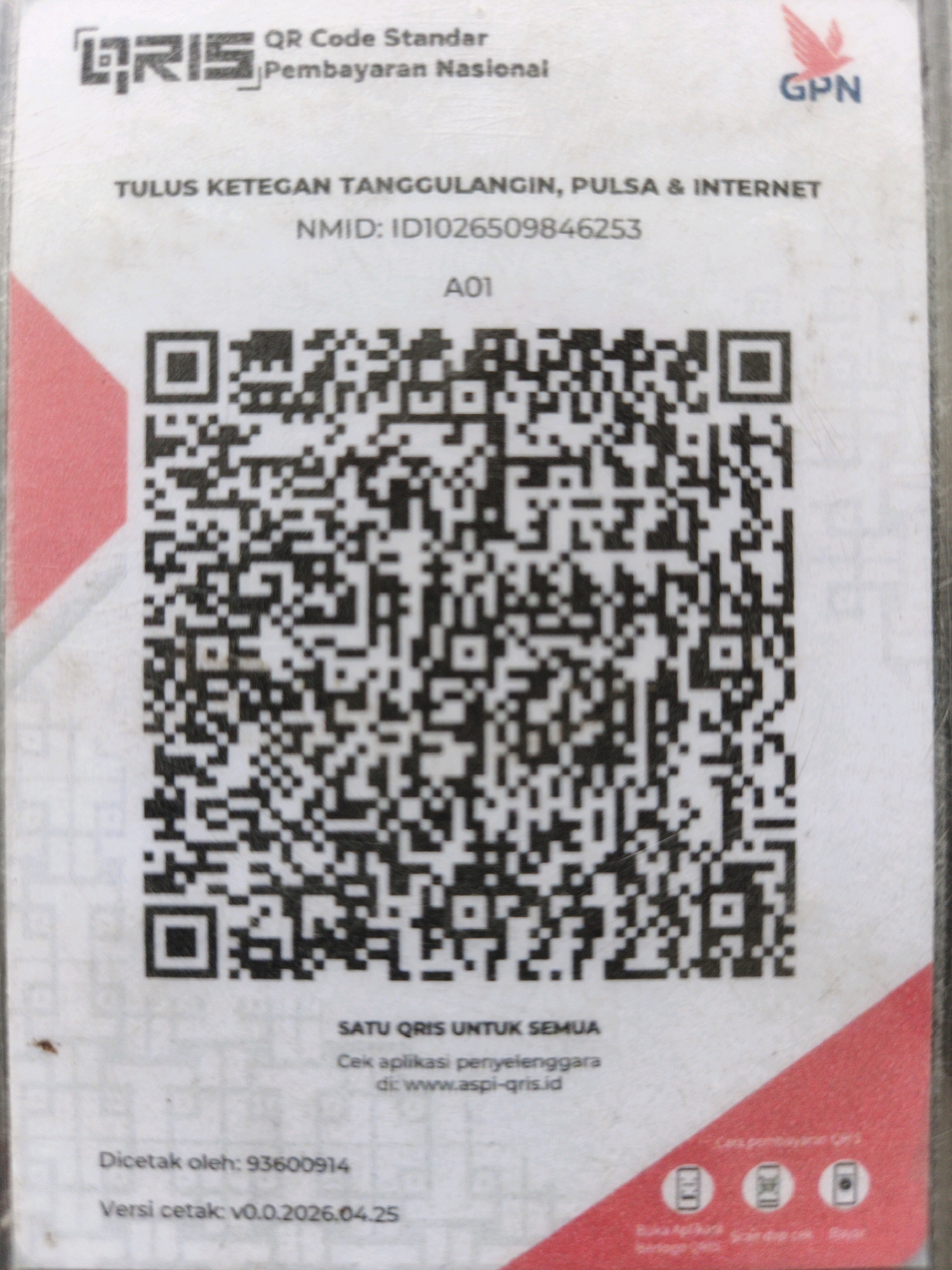 The image is a close-up of a QR code payment sticker. At the top left, the logo "QRIS" is visible, followed by "QR Code Standar Pembayaran Nasional." To the right, a stylized red bird logo and "GPN" are displayed. Below this, text reads "TULUS KETEGAN TANGGULANGIN, PULSA & INTERNET" and "NMID: ID1026509846253" with "A01" below that. The central and largest part of the image is dominated by a black and white QR code. At the bottom, it states "SATU QRIS UNTUK SEMUA," followed by instructions for checking the application at "www.aspi-qris.id." Further down, it indicates "Dicetak oleh: 93600914" and "Versi cetak: v0.0.2026.04.25." The sticker is partially covered by a red triangular graphic on the left and right sides. The background appears to be a textured surface, possibly a wall or counter. There are no people or animals visible in the image. The environment is an indoor setting, suggested by the lighting and the nature of the object. The presence of "Ketegan" in the text indicates the payment system is associated with that location in Indonesia.