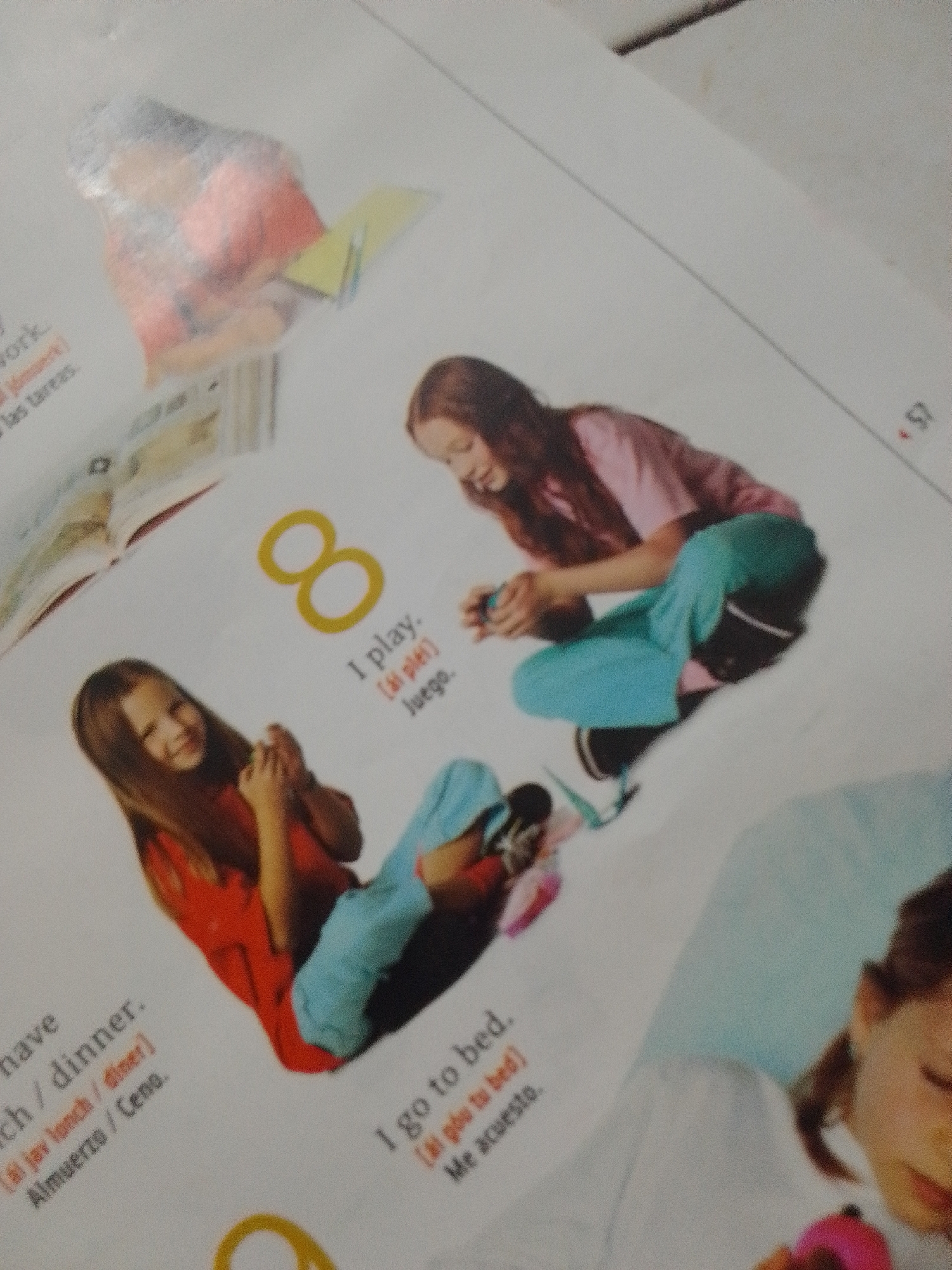 This is a close-up, angled photograph of an open book. The page features illustrations of children and text in English and Spanish. In the upper left, a child in a red shirt is partially obscured, appearing to be reading or working on something yellow. The main focus is on two girls. One girl, with long brown hair, wearing a pink shirt and teal pants, is crouching and holding a small object. To her left, a girl with shoulder-length brown hair, wearing a red shirt and teal pants, is also crouching and smiling while holding an object. The number "8" is prominently displayed in yellow, with the text "I play." and its Spanish translation below. Other text, including "lunch / dinner" and "I go to bed," is visible in smaller font, along with Spanish translations. The background is a plain white surface, likely the book's page. There is no visible indication of the location being La Tagua, Venezuela.