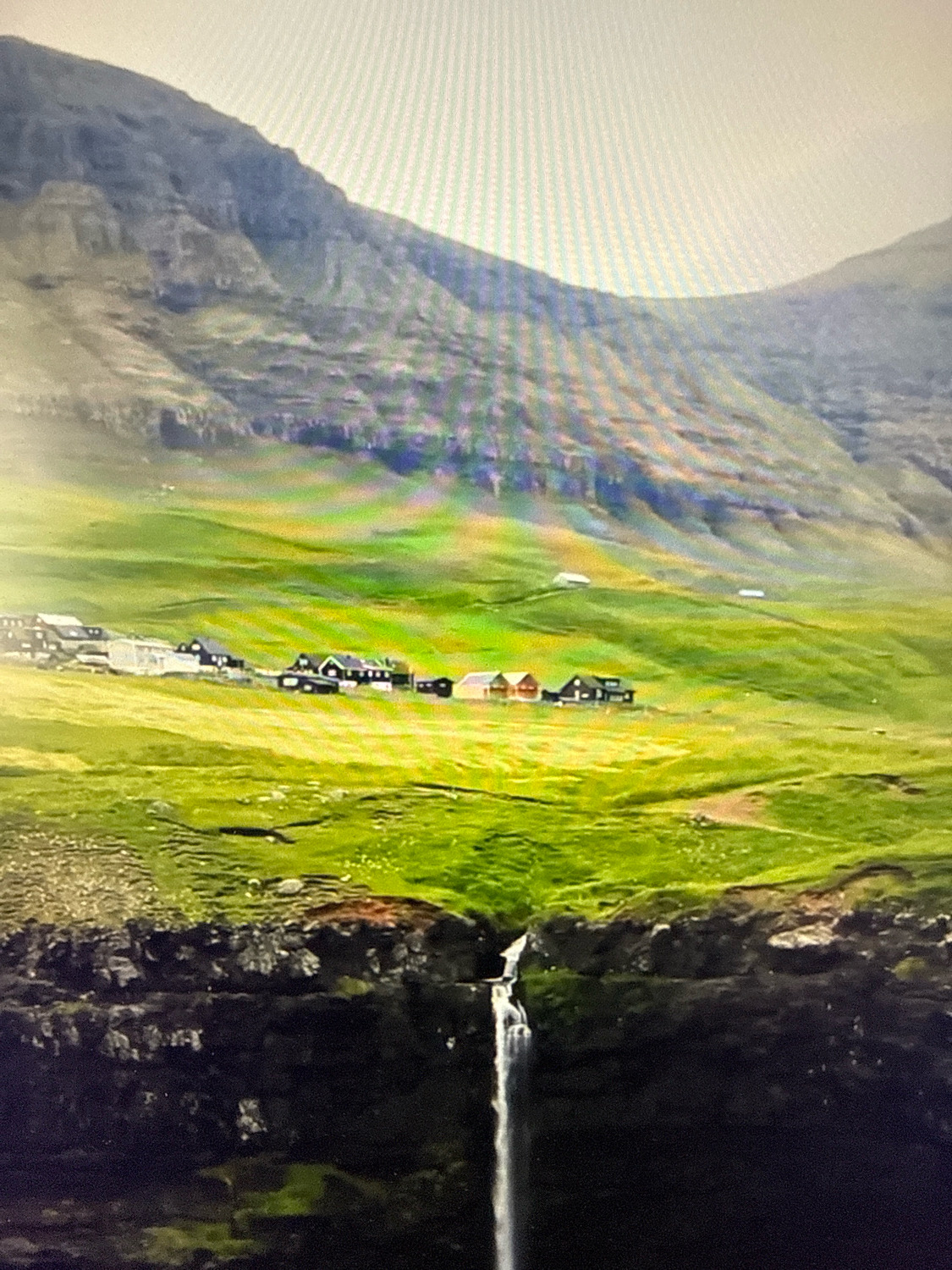The image depicts a picturesque landscape featuring a village nestled on a vibrant green hillside, with a cascading waterfall in the foreground. The village consists of several houses with colorful roofs, including red and orange, and white buildings. The hillside is characterized by lush grass and appears to be part of a mountainous terrain, with a large, textured mountain range dominating the background. The sky is overcast. This scene is not located in Netanya, Israel, but rather appears to be a rural or coastal setting in a location with significant elevation changes and verdant vegetation, possibly resembling the Faroe Islands or a similar European coastal region.