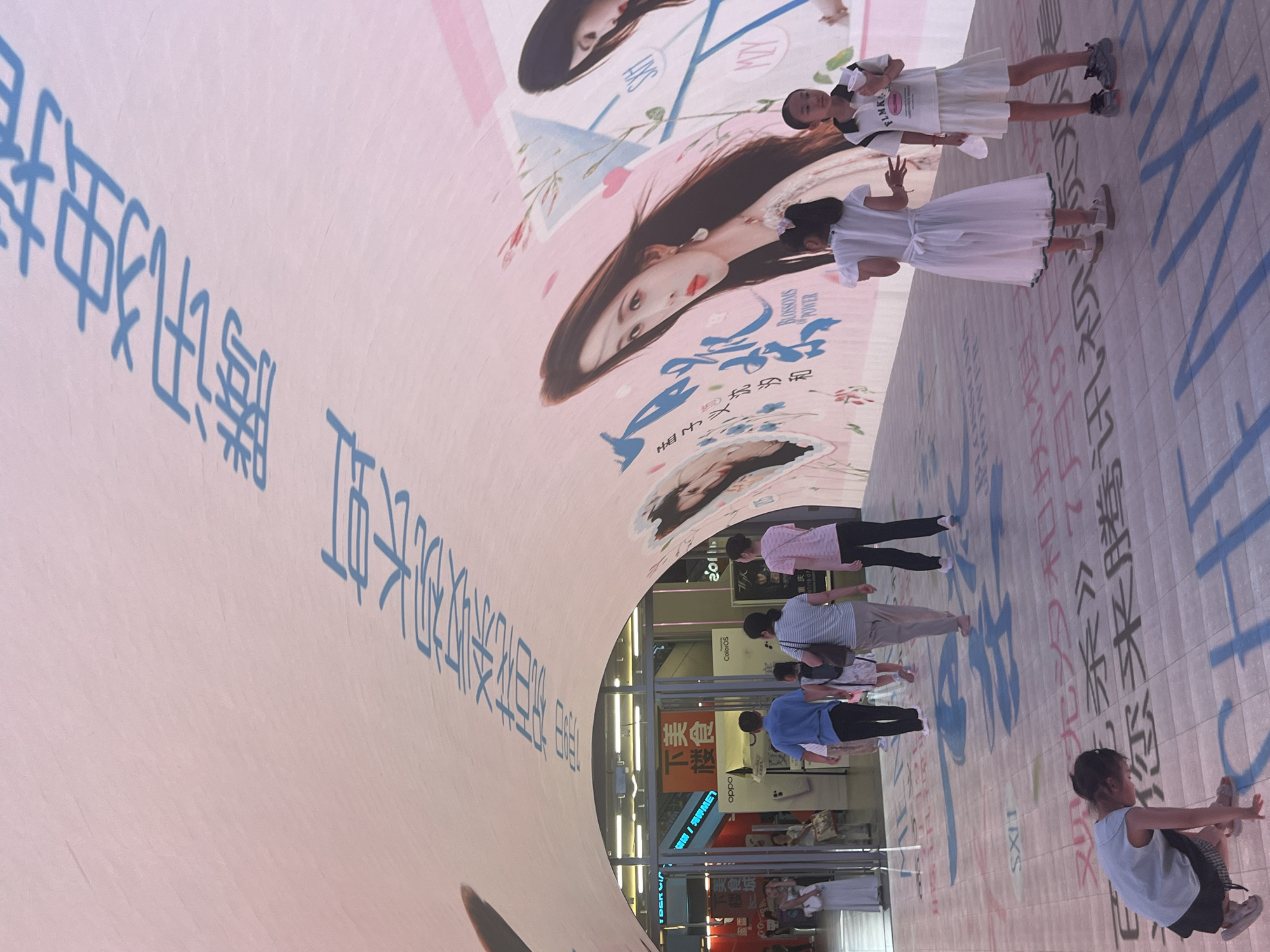 In Yuzhong District, China, a large arched walkway is illuminated by a massive projection of blue Chinese characters across its curved surface and a large image of a woman with dark hair. Beneath this projection, several people are walking through the illuminated space. In the foreground, a young girl in a light blue tank top and checkered skirt is crouching on the ground, looking down. To her right, two girls in white dresses are standing, one with her hands outstretched and the other holding a cup. Further back, a family of four, including two adults and two children, are walking away from the camera. The walkway is paved with square tiles, and more blue Chinese characters are projected onto the floor, creating a vibrant and immersive environment. Shops and escalators are visible through an archway in the background, suggesting a commercial area.