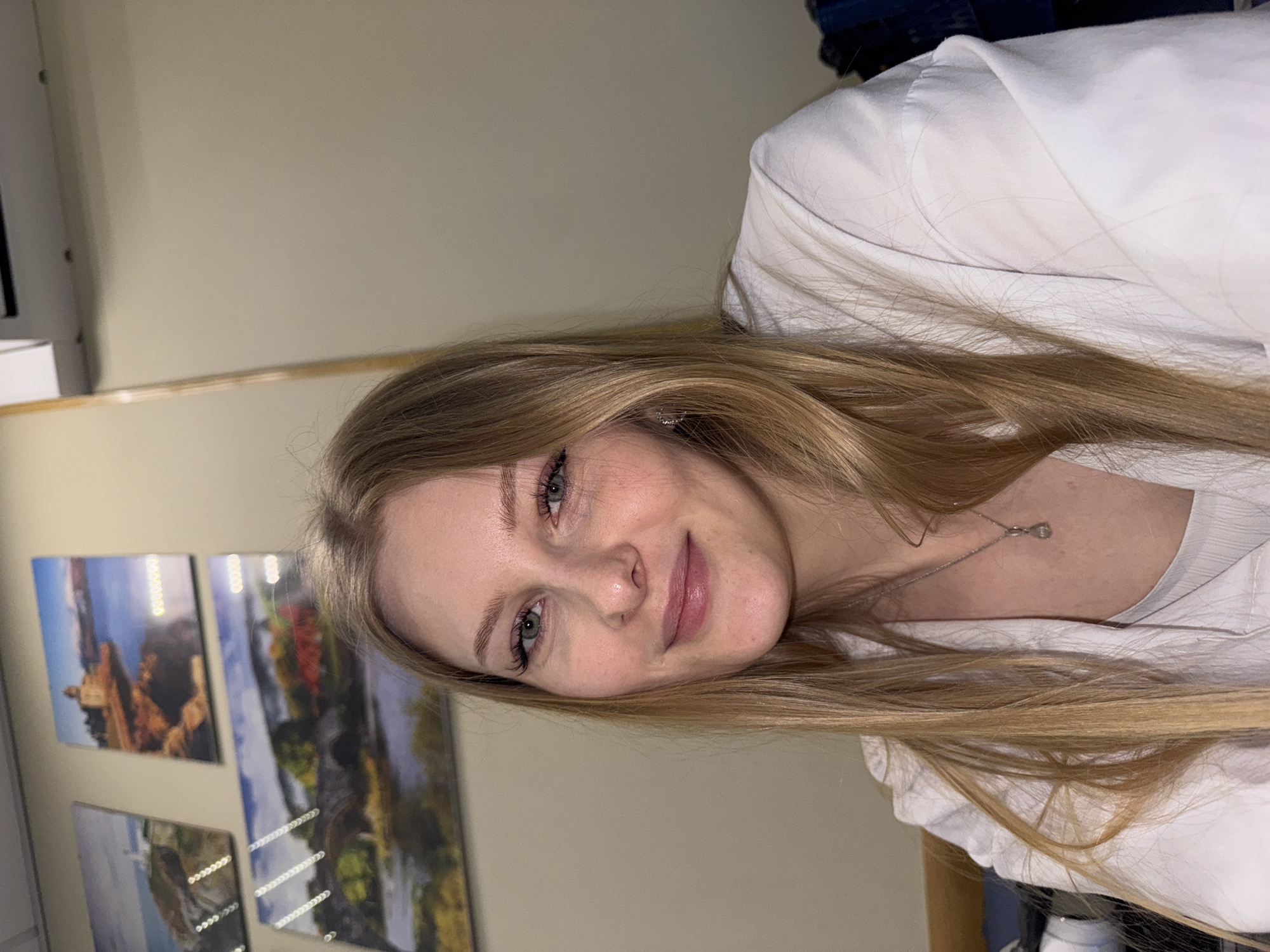 A person with blonde hair and fair skin is facing the camera. She is wearing a white top and a silver necklace. In the background, there are three framed pictures hanging on a beige wall. The pictures appear to be landscape scenes. There is no indication that the image was taken in Perm, Russia.
