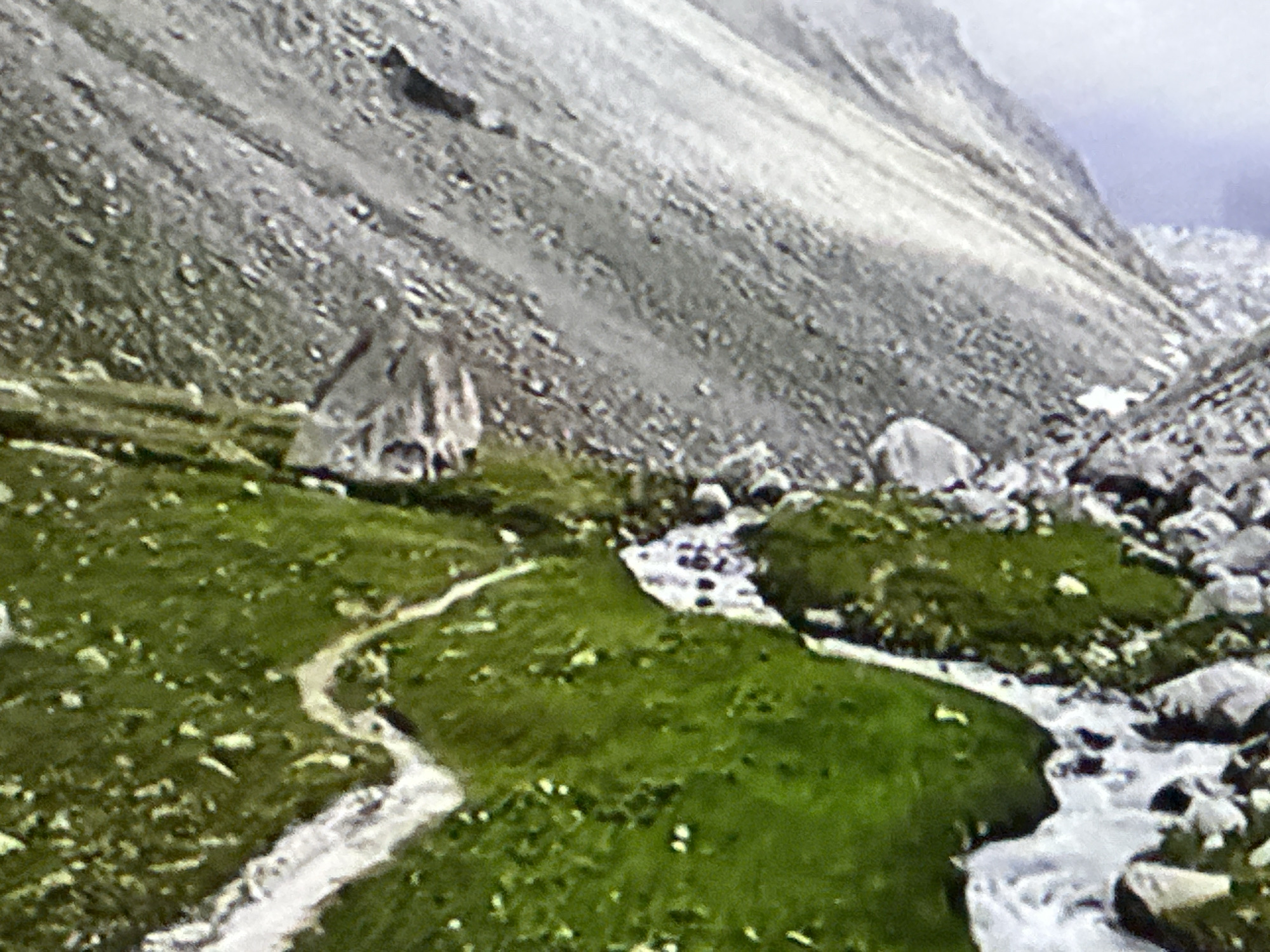The image depicts a mountainous landscape with a winding dirt path leading through a green, grassy area. A stream flows through the scene, bordered by rocks. In the background, a steep, rocky slope rises towards a hazy sky. There are no people or recognizable man-made structures in the image. The location is not Podolsk, Russia, as the scene is clearly a natural mountain environment.