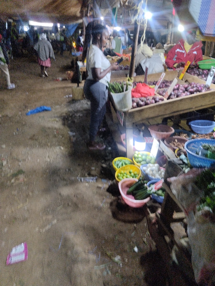 A woman with a ponytail stands at a market stall examining her phone. She is dressed in a white shirt and dark pants. On the table next to her, a variety of produce including onions and leafy greens are displayed. Below the table are baskets filled with cucumbers, limes and garlic. The market is crowded, with other people visible in the background. The ground is uneven and littered with debris. Overhead lights illuminate the scene.