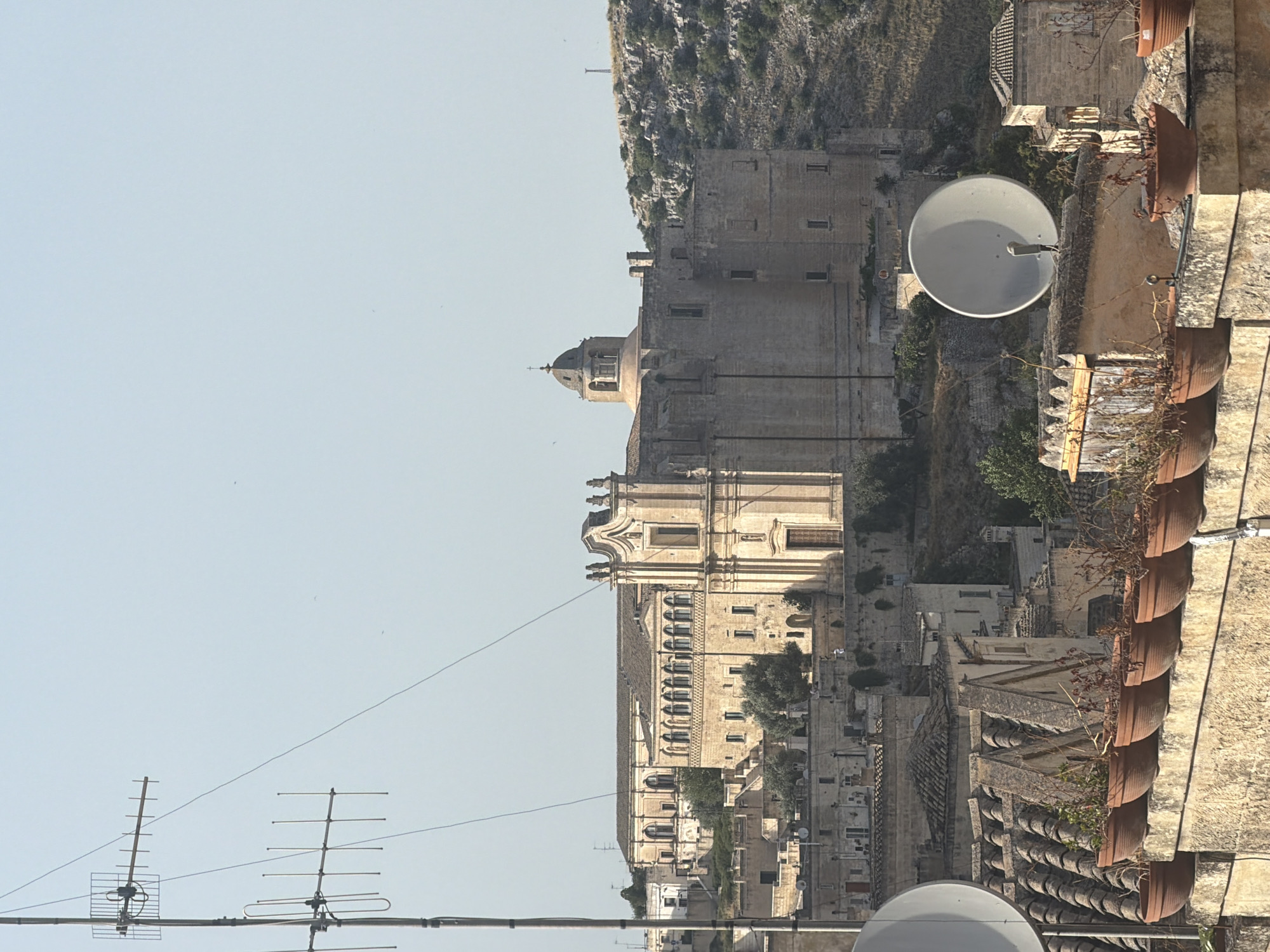 The image depicts a view of the city of Matera, Italy, featuring the historic Chiesa di San Francesco da Paola on the left and the Chiesa di San Pietro Caveoso on the right, nestled within the Sassi districts. In the foreground, a stone wall is lined with several terracotta pots containing dry plants. Two satellite dishes are visible, one larger and positioned centrally, and another smaller one on the left. An antenna array stands tall on the left side of the frame. The background consists of the characteristic stone buildings of Matera, carved into the rocky landscape, with a clear, pale blue sky overhead. The overall scene conveys a sense of historical depth and a dry, sun-drenched atmosphere. There are no people visible in the image.