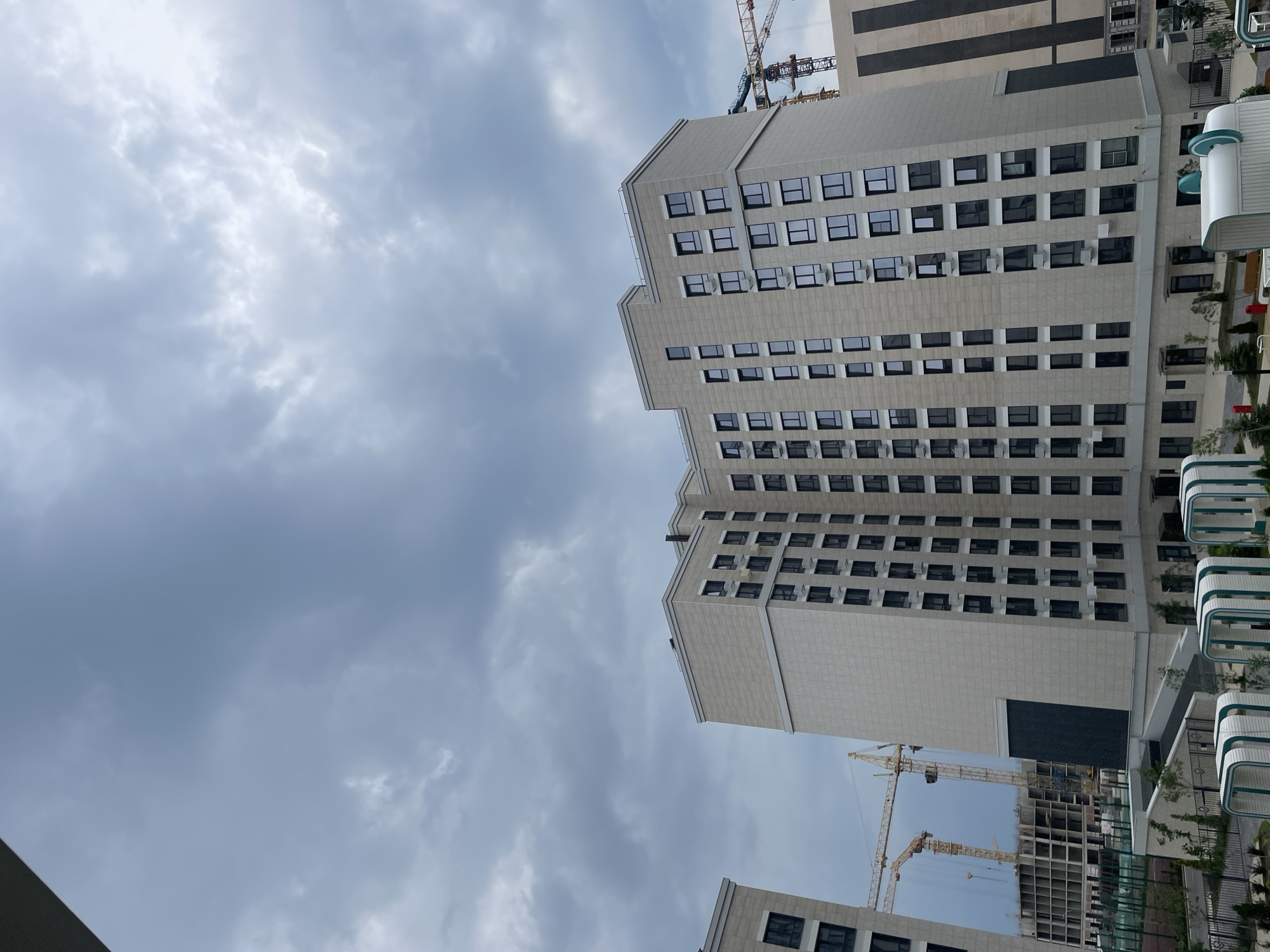 This media file captures a view of a modern residential building under construction, set against a cloudy sky. The building features a light-colored facade with numerous windows arranged in a grid pattern. Some windows are open, suggesting ongoing construction or habitability. Several air conditioning units are visible beneath some of the windows.
The scene includes construction cranes, indicating that the building is not yet fully completed. In the background, other buildings and structures are partially visible. The sky is overcast, suggesting a cloudy or possibly impending rain. The overall setting appears to be an urban or suburban environment, given the presence of multi-story buildings and construction activity. There is no discernible text within the image. The orientation of the image is such that the buildings are viewed from a low angle, looking upwards. The lighting suggests it is daytime, but the cloud cover creates a subdued atmosphere.