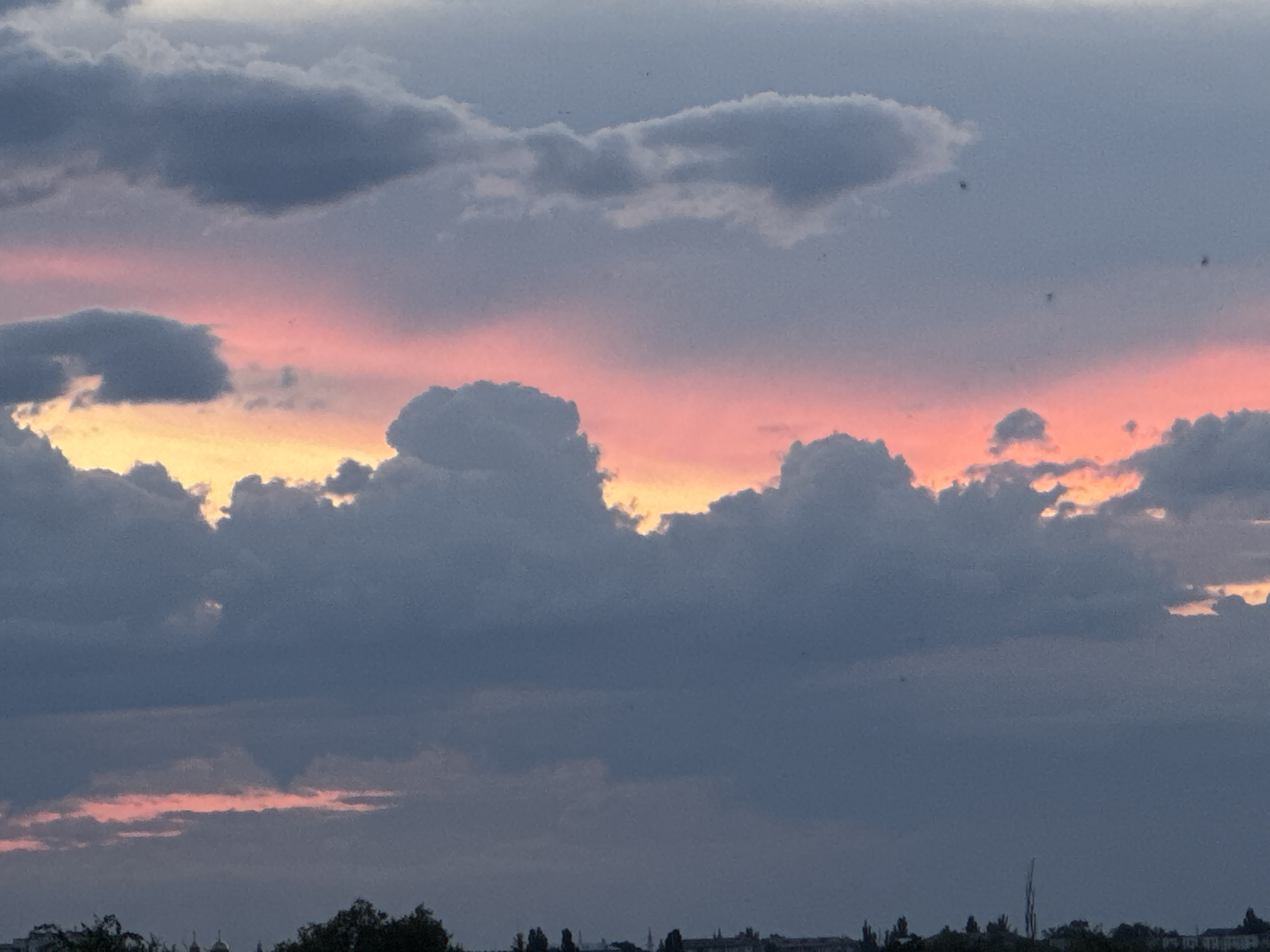 The image depicts a cloudy sky at dusk, with hints of pink and yellow light from the setting sun reflecting off the undersides of the clouds. The clouds are predominantly gray and dark, with some lighter, more textured formations. In the lower portion of the image, a silhouetted treeline and the faint outlines of buildings are visible, suggesting an urban or suburban landscape. It is not possible to definitively confirm the location as Tiraspol, Moldova based solely on the provided image. There are no people, specific objects, or identifiable actions captured in the scene.
