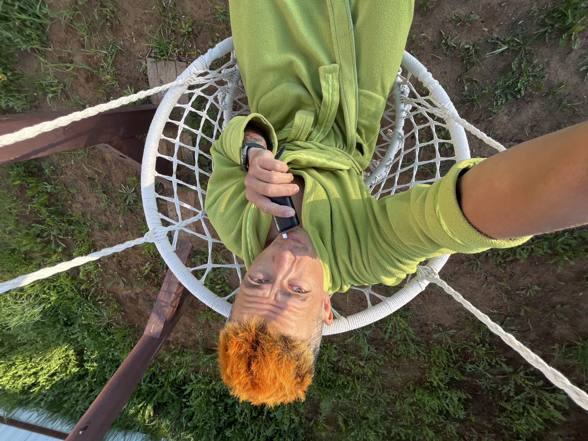 A middle-aged person with orange hair is reclined in a white hammock-style chair, wearing a light green robe. The person is holding a black object up to their mouth, possibly a vaping device. The chair is suspended from a wooden frame with thick white ropes. The background consists of grass and dirt. The image appears to have been taken from directly above. There is no information to verify the location in Chaikovsky, Russia.