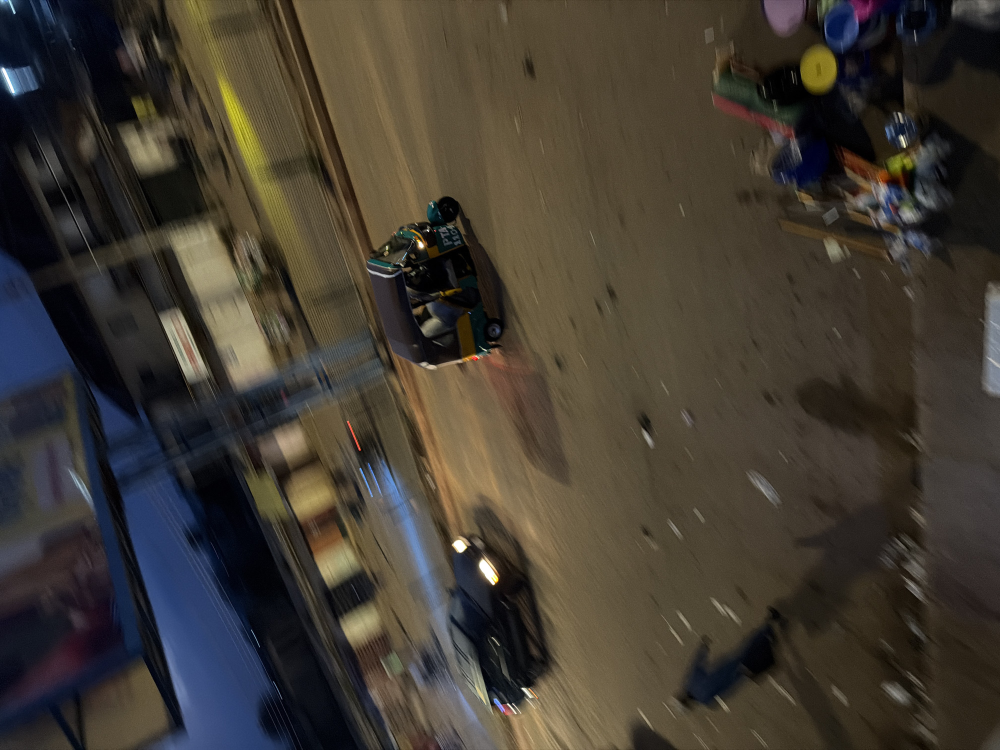 The image shows a street scene at night in Bukuru, Nigeria. In the foreground, a blue and yellow auto-rickshaw, labeled "PTA 1204," is moving to the right, its headlights illuminated. To its left, a dark-colored car is also in motion, with its headlights and taillights visible. In the lower left quadrant, a person in a dark blue garment appears to be walking or running away from the viewer, casting a long shadow. The street is littered with debris. In the background, buildings and streetlights are visible, though blurred due to motion. The overall impression is of a busy, moving street scene at dusk or night.