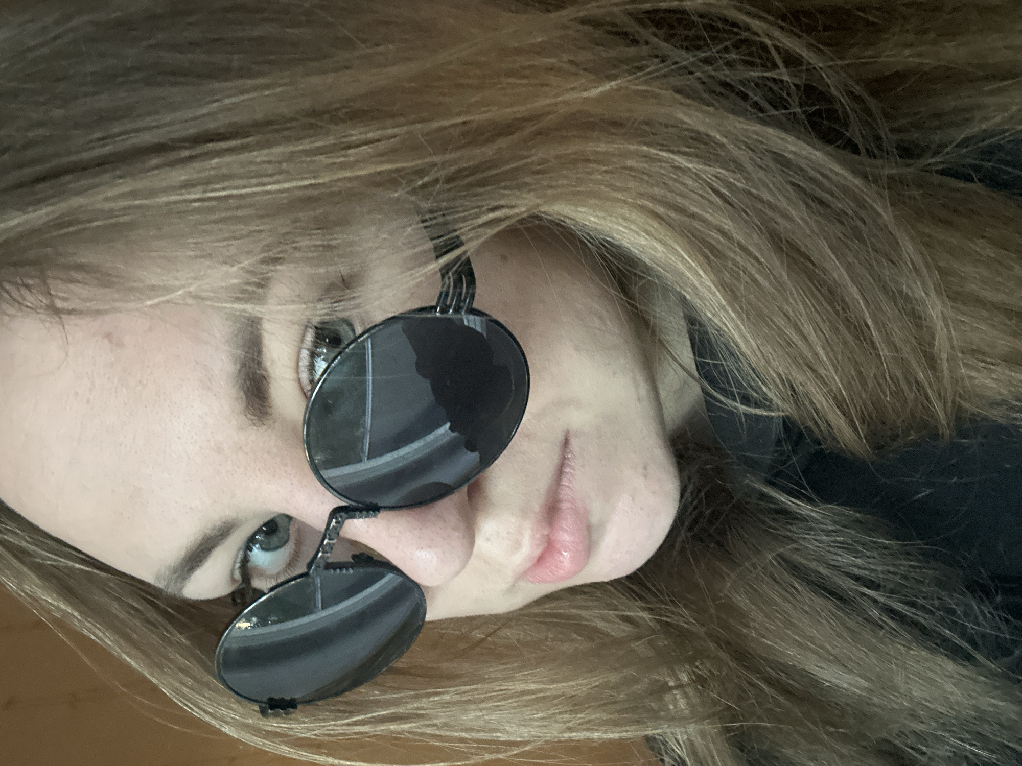 A close-up, angled portrait features a person with long, flowing light brown hair with blonde highlights. The individual is wearing round, dark-tinted sunglasses that are perched on their nose, partially obscuring their eyes. One eye, appearing light-colored (possibly blue or green), is visible above the rim of the sunglasses and looks slightly upwards. The other eye is mostly hidden behind the left lens. The person's lips are partially visible, light pink, and slightly parted. Their skin tone appears fair. The hair cascades around the face, filling much of the background, with some dark fabric (possibly clothing) visible in the deeper parts of the hair. The overall perspective suggests the person is either lying down and looking up at the camera or the image is heavily rotated. No specific location cues, time of day, or visible text are discernible. The expression appears neutral or contemplative.