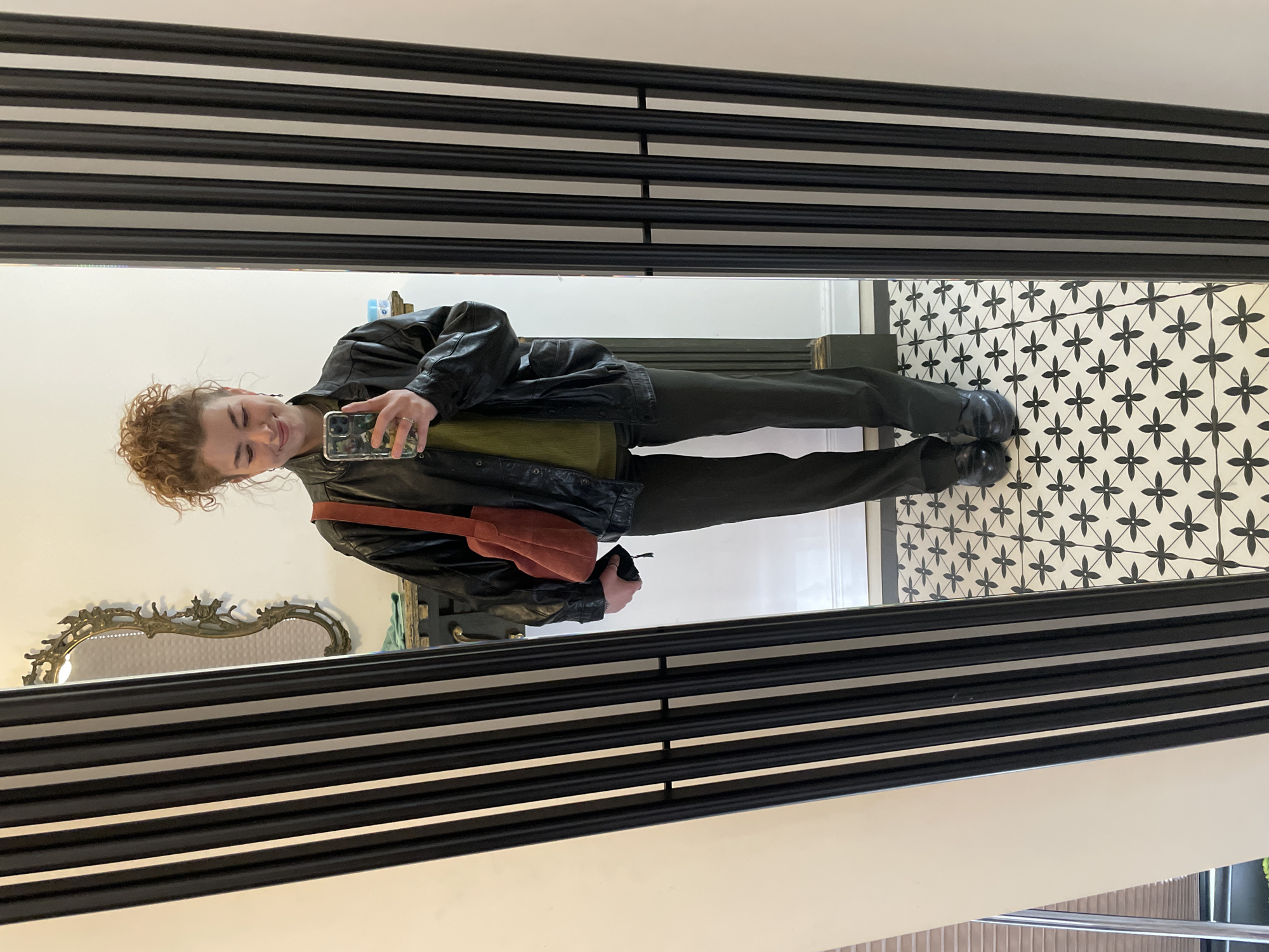 The image shows a young woman taking a selfie in a mirror. She is wearing a black leather jacket, dark pants, and black shoes. She has a green top and a red bag. The background shows a patterned floor and decorative elements, including a mirror and a striped frame. The location is likely a building interior in Tbilisi, Georgia, given the architecture and style of the decorations.
