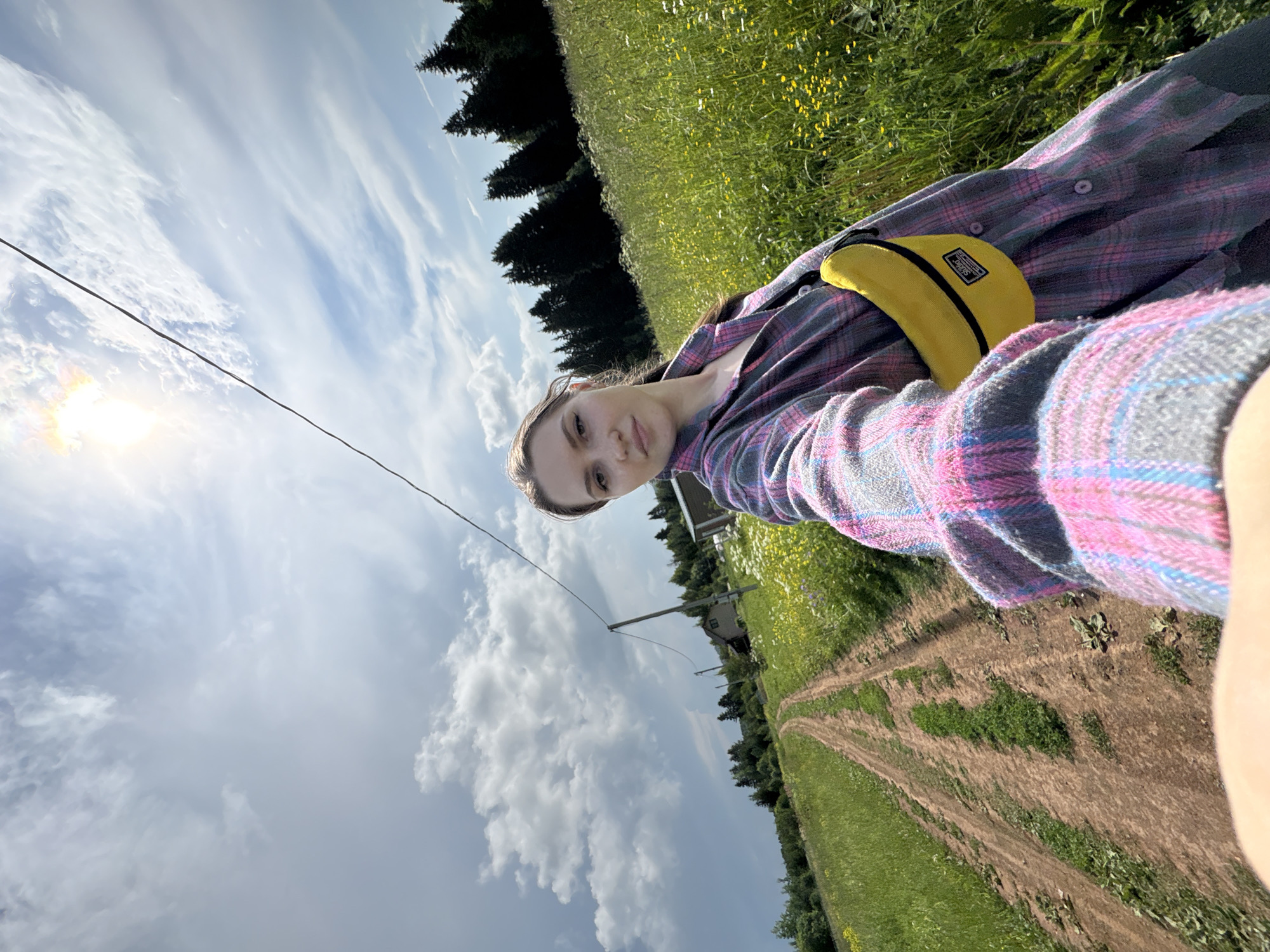 A young woman, with long brown hair and fair skin, is taking a selfie in a rural landscape. She is wearing a pink, purple, and grey plaid shirt and a bright yellow fanny pack. She is standing in a field of tall green grass dotted with small yellow flowers. A dirt road stretches out behind her, leading towards a line of dark green evergreen trees. In the distance, a small house and a utility pole are visible. The sky is a bright blue with scattered white clouds, and the sun is shining brightly. The scene appears to be set in the countryside, possibly near the village of Kumyonys in Russia, given the context provided.