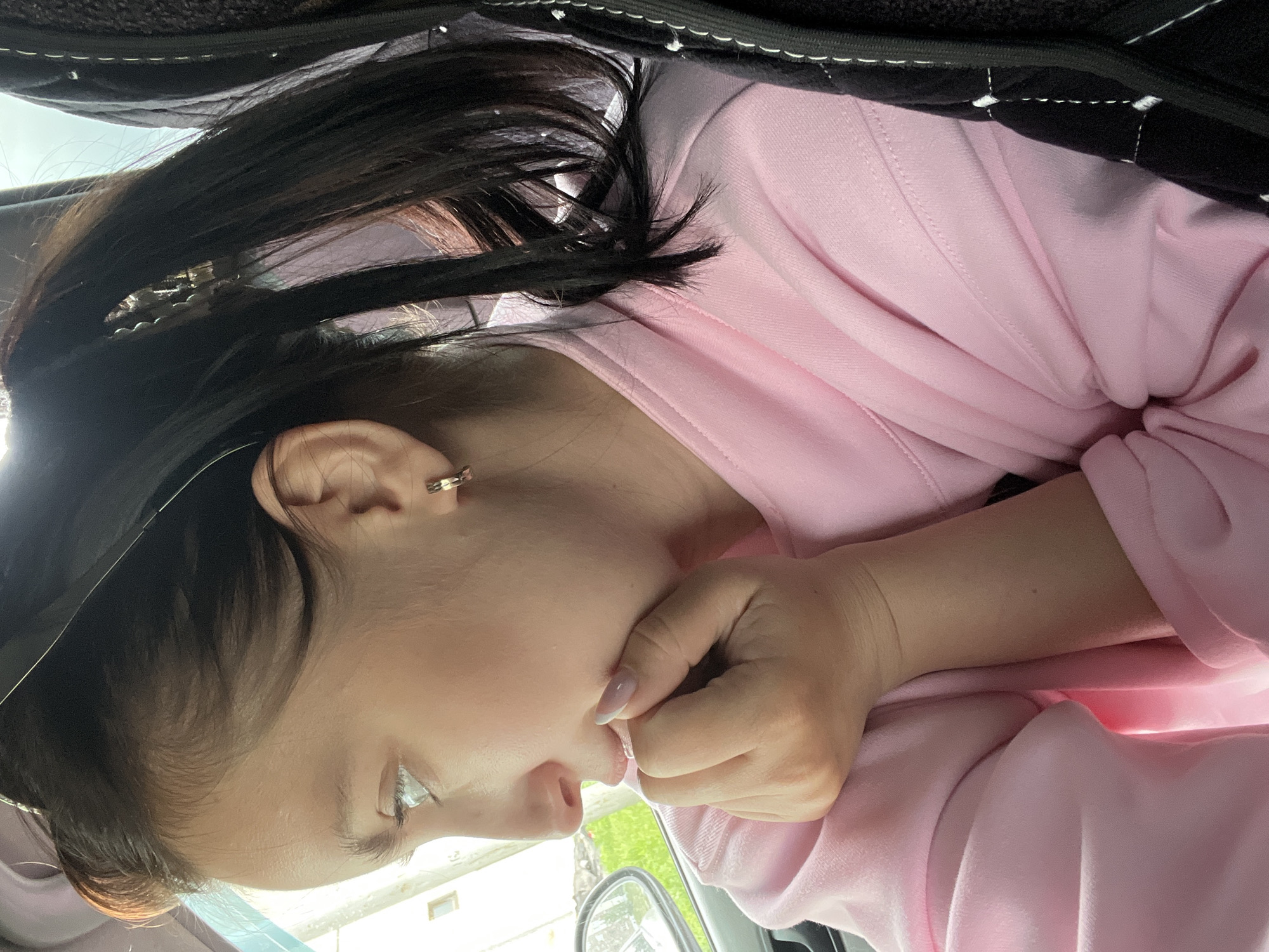 A female subject is depicted in a close-up, profile view, positioned within a vehicle interior. Her gaze is directed towards the left, out a window. She has dark brown hair, which is pulled back and secured with a light-colored hair clip, forming a ponytail. A small, gold-toned hoop earring is visible in her right ear. Her complexion is fair. She is wearing a light pink, long-sleeved garment, consistent with a sweatshirt or hoodie, made of a soft fabric. Her right hand is placed beneath her chin, with fingers curled; her fingernails are shaped and have a light pink base with white tips. Through the vehicle's window, a partial view of a light-colored building with a square window and green foliage is visible. A portion of the car's side mirror also reflects green vegetation. On the far right, a dark, possibly quilted or textured surface, characteristic of a car seat or headrest, is present. The scene is illuminated by natural light, suggesting daytime. The location is the city of Markovskiy, Russia.