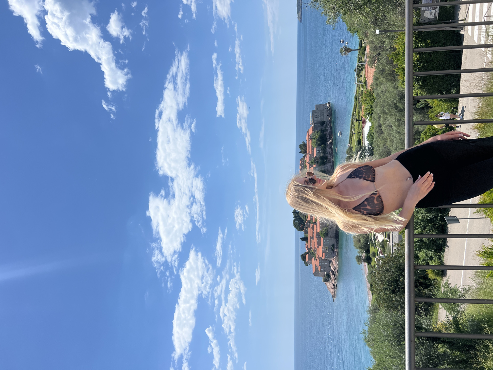 The image shows a woman in a bikini and black leggings leaning against a railing with a view of Sveti Stefan, Montenegro. The sea stretches out to the island town, and the sky is bright blue with scattered white clouds. The railing is gray and runs horizontally across the lower right side of the frame. A road can be seen through the greenery behind the railing. The image is rotated ninety degrees counter-clockwise.