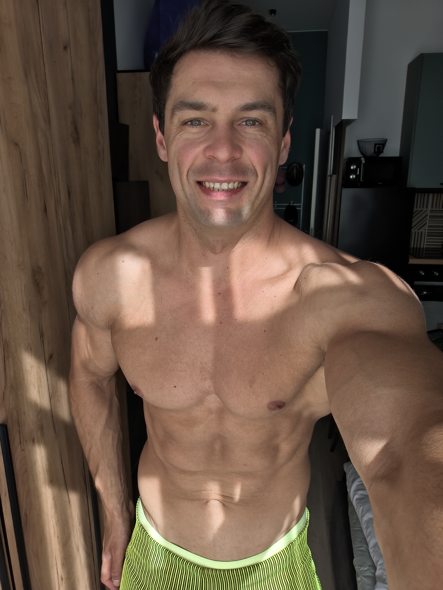 This image, taken in Moscow, Russia, features a shirtless adult male posing for a photograph in an indoor residential setting, likely an apartment or studio. The man, with short dark brown hair and greenish-brown eyes, smiles broadly towards the camera, displaying a muscular and lean physique with defined chest, shoulders, arms, and visible abdominal muscles. He wears neon yellow-green striped athletic shorts. Strong natural sunlight from an unseen window to the left casts distinct rectangular shadows across his chest and the wooden paneling in the background.
To the left of the subject is light-colored wooden paneling, possibly a door or closet. The right side of the background reveals a compact kitchen area with dark lower cabinetry, a black microwave, a grey bowl, and a dark refrigerator. Above these are teal/mint green overhead cabinets and a geometric black and white tiled backsplash. A dark blue/grey wall is visible in the upper right, and crumpled white fabric, possibly bedding, is partially visible in the lower right corner.