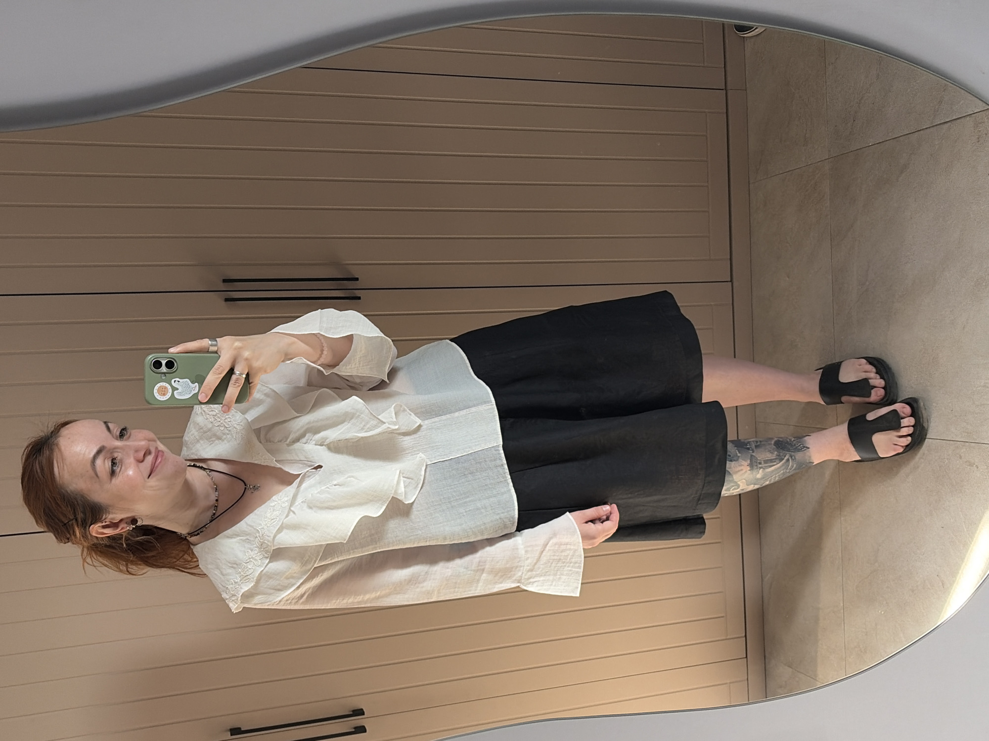 The image shows a mirror selfie of a fair-skinned woman with auburn hair. She is smiling and holding an olive-green smartphone in her right hand, its camera facing the mirror. She is wearing a white, long-sleeved, ruffled blouse and black culottes. The woman is also wearing a necklace and black flip-flops. Her left calf is visibly tattooed. The reflection reveals she is standing in front of what appears to be a light brown cabinet or wardrobe. The floor is covered in neutral-toned square tiles. There is no verifiable information about the location being Chaikovsky, Russia.