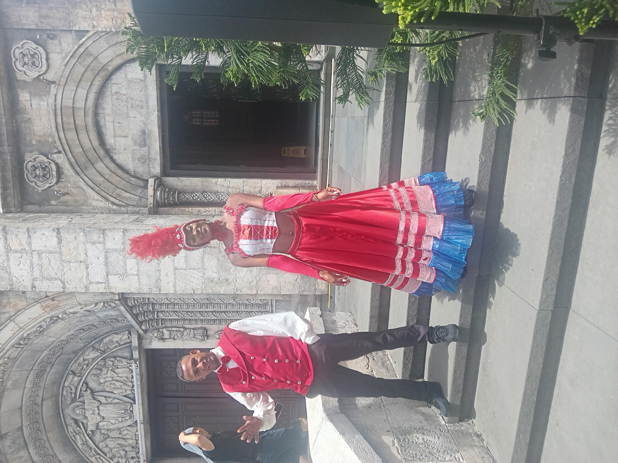 Two performers, a man and a woman, are posing on the steps of a stone building. The man, wearing a red vest over a white shirt and a red bowtie, stands on the left, with his left hand in his pocket and his right hand raised as if waving. He is wearing black pants and dark sneakers. The woman, on the right, is dressed in a red and white costume with a feathered headdress. Her costume includes a ruffled top, a long, layered red skirt with blue trim, and long red sleeves. She has a red bandana around her neck. Behind them, the stone facade of a building features arched doorways and decorative carvings. A lush green tree branch is visible on the right side of the frame. The scene appears to be in an outdoor, possibly tourist, location. This building is identified as the D'Jungle Bar area within Sun World Ba Na Hills, in the city of Đà Nẵng, Vietnam.