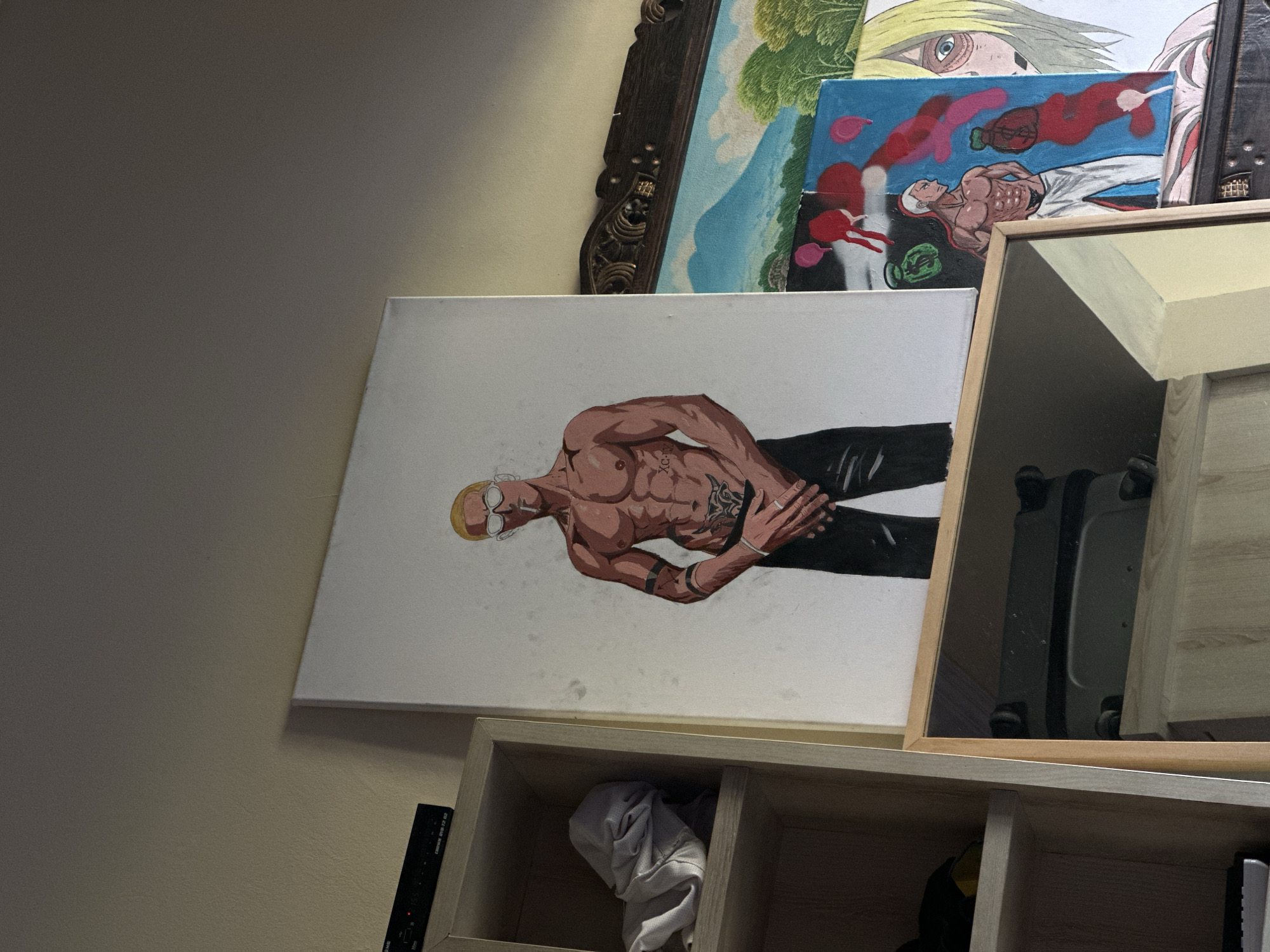 A painting of a muscular man with blond hair and sunglasses, smoking a cigarette and wearing black pants and a silver ring, is displayed on a white canvas. The painting is placed on a shelf next to a wooden bookshelf containing a gray crumpled garment. To the right of the central painting, another canvas features a drawing of a muscular man with white hair and red accents, standing next to a larger artwork depicting a landscape with mountains and trees, and a character with blond hair and blue eyes. The scene is indoors, with the paintings leaning against a wall.