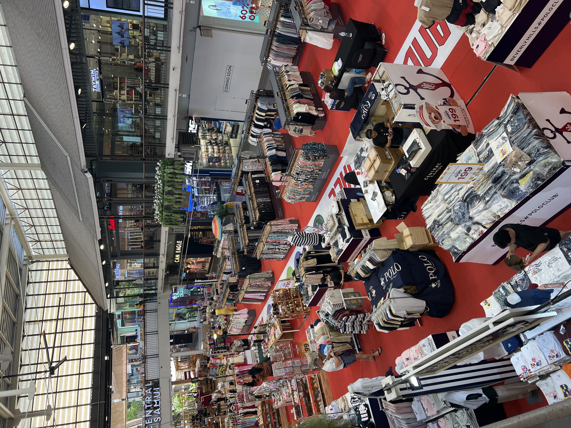 This is a high-angle, slightly tilted shot of a busy shopping mall. The scene is dominated by displays of clothing, primarily t-shirts and polo shirts, arranged on racks and tables on a vibrant red carpet. Several people are browsing the merchandise, some are attending to the displays, and one person appears to be a vendor or staff member.
Prominent text visible includes brand names like "POLO CLUB," "BEVERLY HILLS," and "CAN EAGLE." There are also signs indicating sales, such as "60% OFF" and price tags with numbers like "199" and "299." A sign reads "COMING SOON," suggesting a new store or section is opening. Another sign says "CENTRAL SAMUI," which, combined with the provided location context of "บ้านบางรักษ์, Thailand," identifies the mall. The lighting suggests it is daytime, likely indoors in a well-lit commercial space. The overall atmosphere appears to be bustling and commercial, with a focus on retail shopping. The arrangement of goods and the presence of shoppers indicate a typical retail environment.