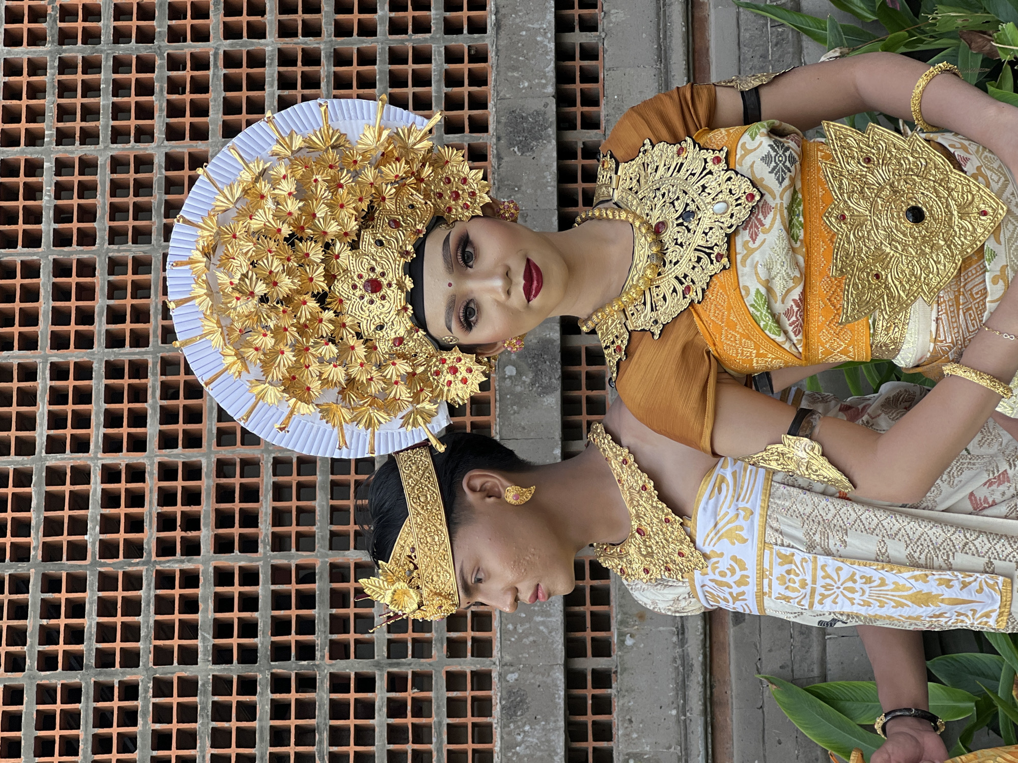 Two individuals in traditional Balinese attire stand in front of a brick wall. The person on the left, wearing a gold crown and elaborate gold necklace, faces left in profile. The person on the right, adorned with a large, ornate gold headdress with white and gold accents, and a matching gold necklace and belt, faces forward. Both are dressed in intricately patterned white and gold garments. The background consists of a wall made of orange bricks with square openings, suggesting an outdoor setting in Denpasar, Indonesia. Green foliage is visible at the bottom of the image.
