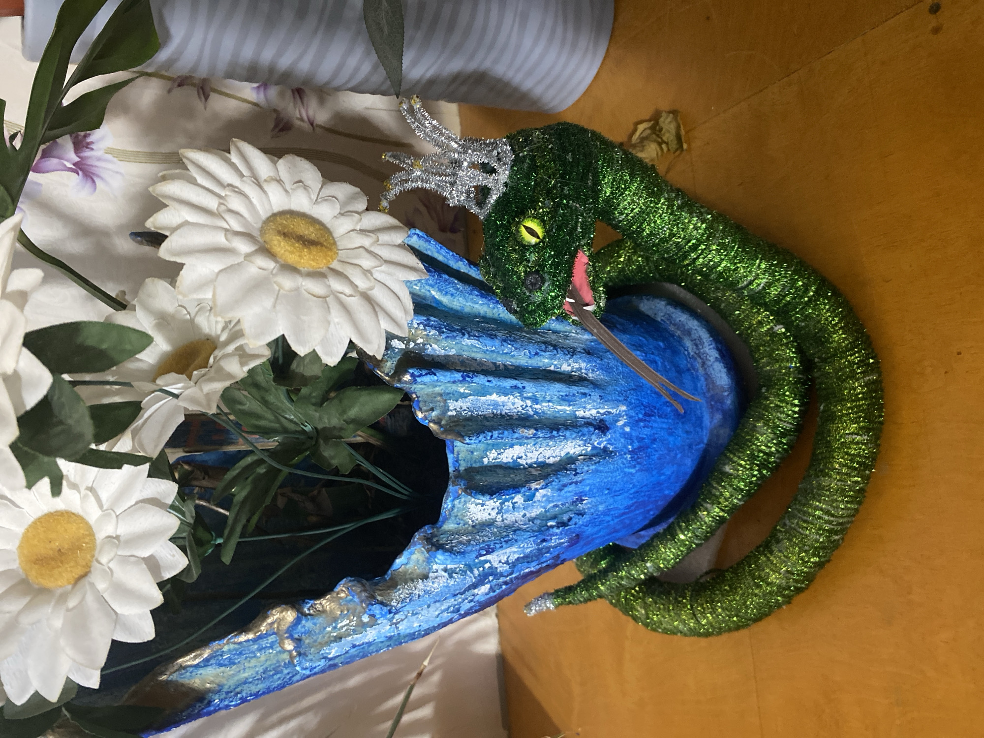 The image depicts an indoor scene in the city of Noviy, Russia. A blue and gold textured, irregularly shaped pot or vase stands on a flat, orange-brown wooden surface. The pot contains multiple artificial white daisy flowers with prominent yellow centers, interspersed with green artificial foliage. Coiled around the base of the pot is a decorative, green glitter-covered snake figurine. The snake features distinct yellow-green eyes with black pupils, a pink tongue protruding from its mouth, and a small, silver glitter crown positioned on its head. In the background, a light-colored fabric with faint purple floral patterns is visible, along with a partial view of a ribbed, light grey cylindrical object to the right. No human subjects are present.