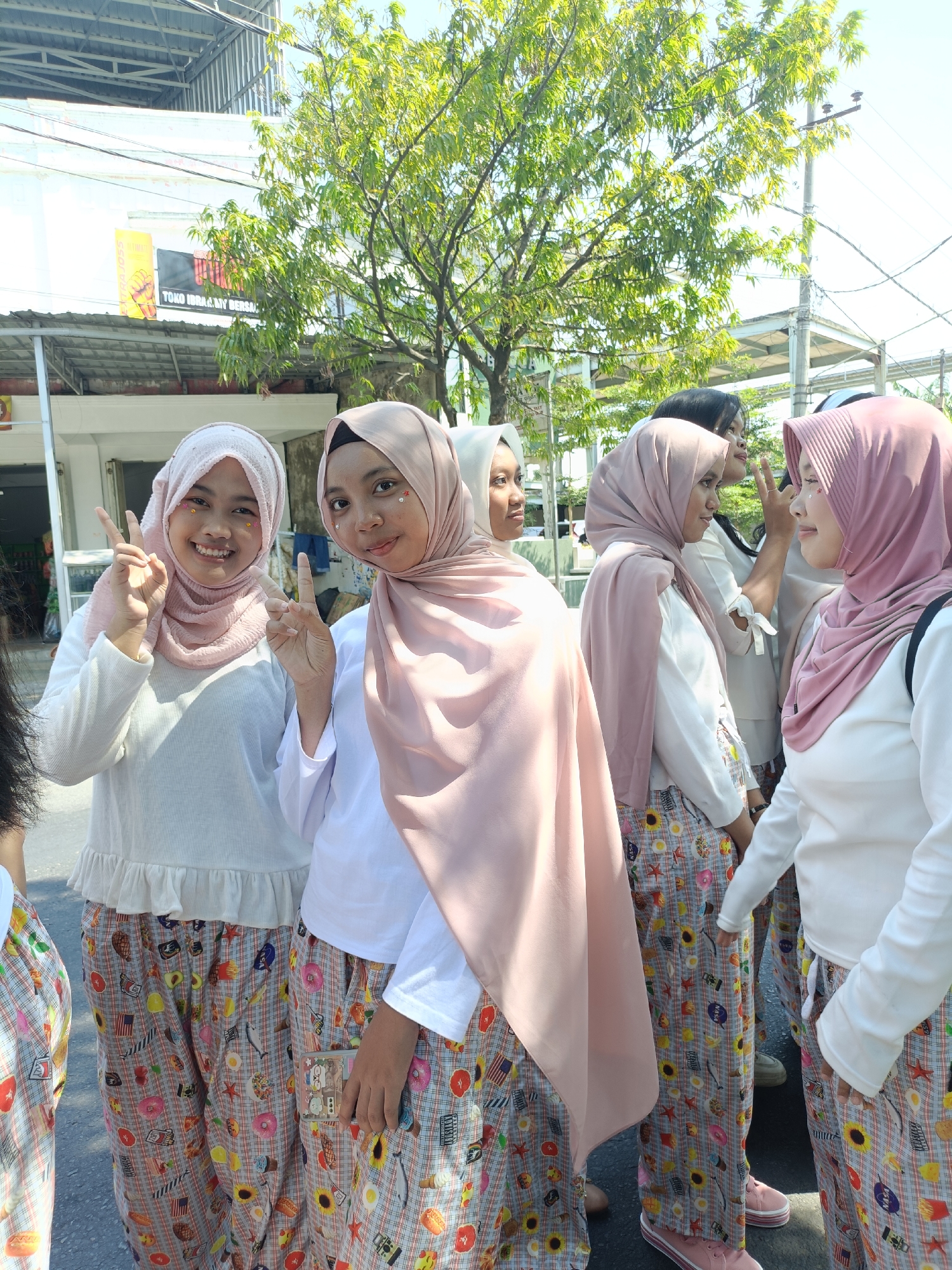 In the city of Kediri, Indonesia, four young women wearing matching plaid pajamas adorned with food motifs and white long-sleeved shirts stand outdoors. They are also wearing pink hijabs. Two of the women are in the foreground, posing for the camera with peace signs and smiles. One has glitter applied to her face. In the background, a store with signage and a tree with green foliage are visible, suggesting an urban or semi-urban setting. The lighting indicates it is daytime, with sunlight casting shadows. The individuals appear to be participating in a coordinated event or activity.