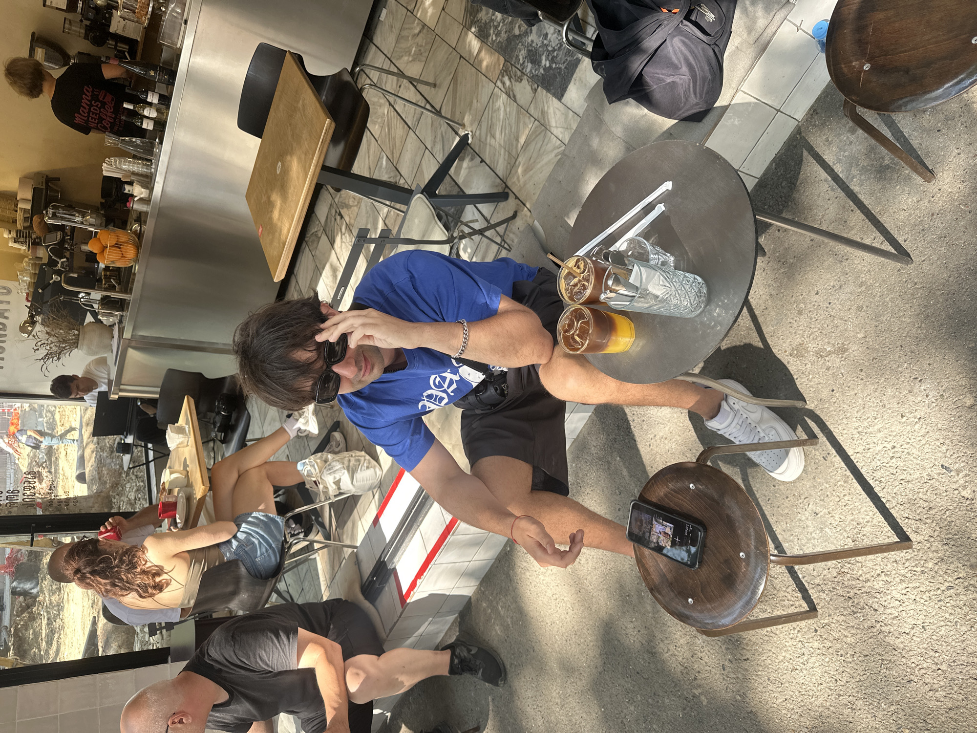 In an outdoor seating area of a cafe in Tbilisi, Georgia, a young man in a blue t-shirt and dark shorts is seated on a concrete step. He is looking to his right, with his left hand touching his sunglasses. In front of him, on the ground, is a small wooden stool with a smartphone on it. Next to the stool is a round metal table with two iced drinks, a glass, and two straws. Further away, a man in a black t-shirt and shorts sits on a bench, and a woman in a denim skirt and a tank top is seated at a table with a coffee cup. In the background, a barista with short hair wearing a black t-shirt with text on the back stands behind a counter with coffee machines and shelves of bottles. The cafe has a modern, industrial aesthetic with metal and wood elements. The sunlight casts shadows on the ground, indicating it is daytime. Construction can be seen in the distance through an open doorway.